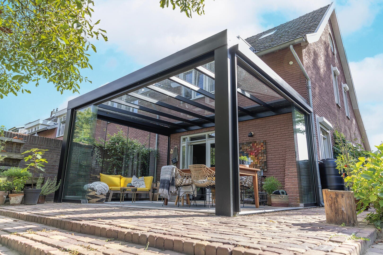 Un abri de terrasse moderne en verre et en métal, adossé à une maison en briques, avec des sièges confortables avec des coussins, une table en bois et des plantes en pot, le tout sous un ciel bleu limpide.
