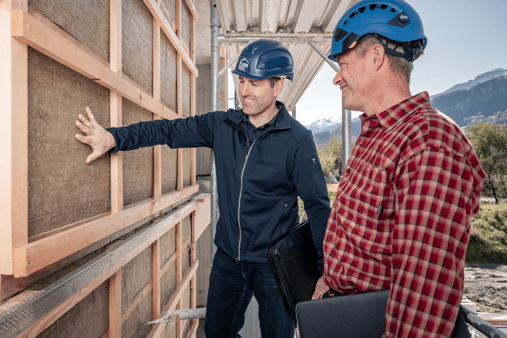 Zwei Männer mit blauen Helmen inspizieren die Außenisolierung eines im Bau befindlichen Gebäudes. Ein Mann gestikuliert in Richtung der Wand, während der andere, in einem karierten Hemd, einen schwarzen Ordner hält. Im Hintergrund sind Berge zu sehen.