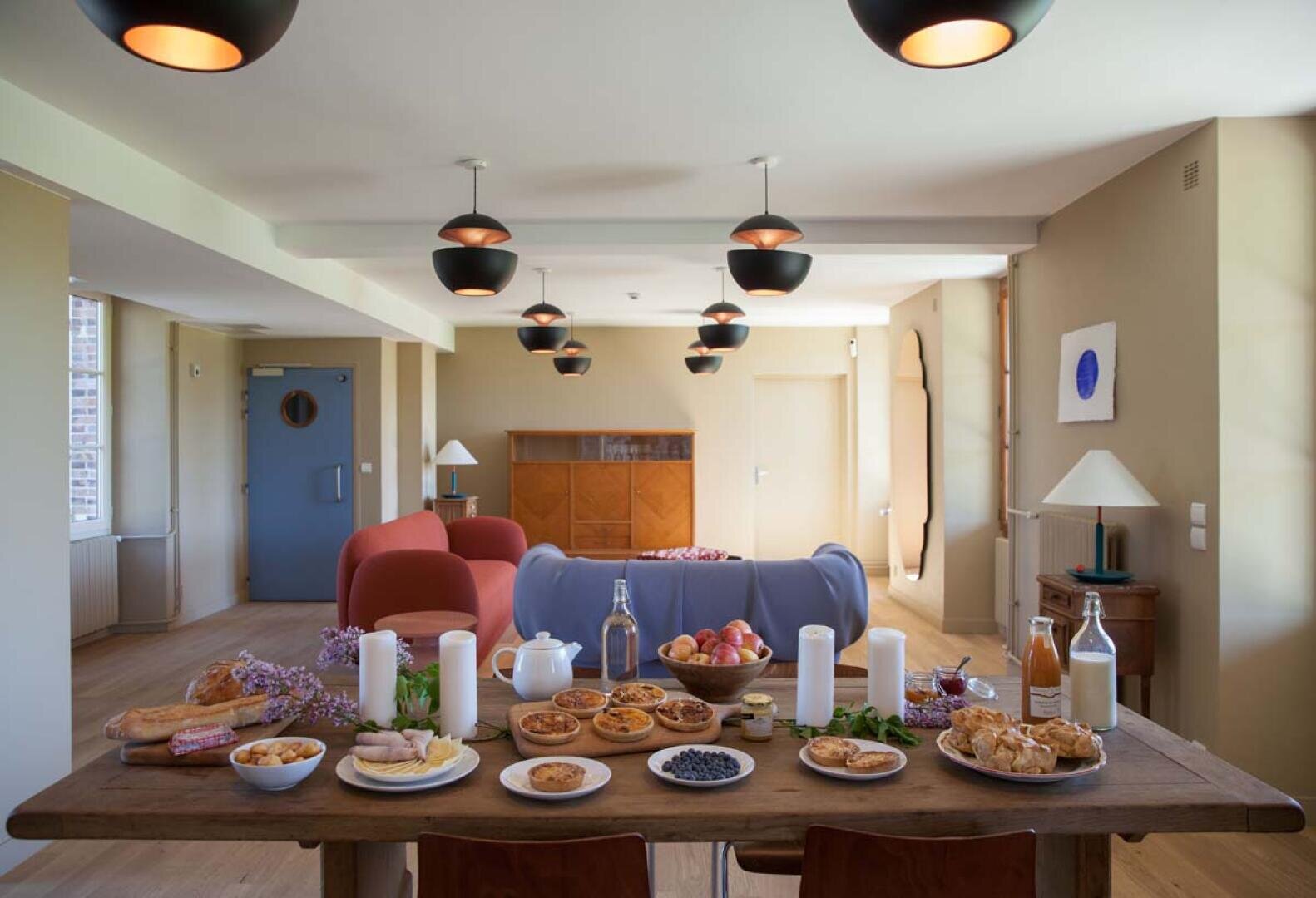 Une table à manger en bois garnie de pâtisseries, de fruits, de pain, de fromage, de boissons et de bougies dans un espace de vie moderne et lumineux avec des canapés, des œuvres d'art murales et des lampes suspendues. La lumière naturelle remplit la pièce.