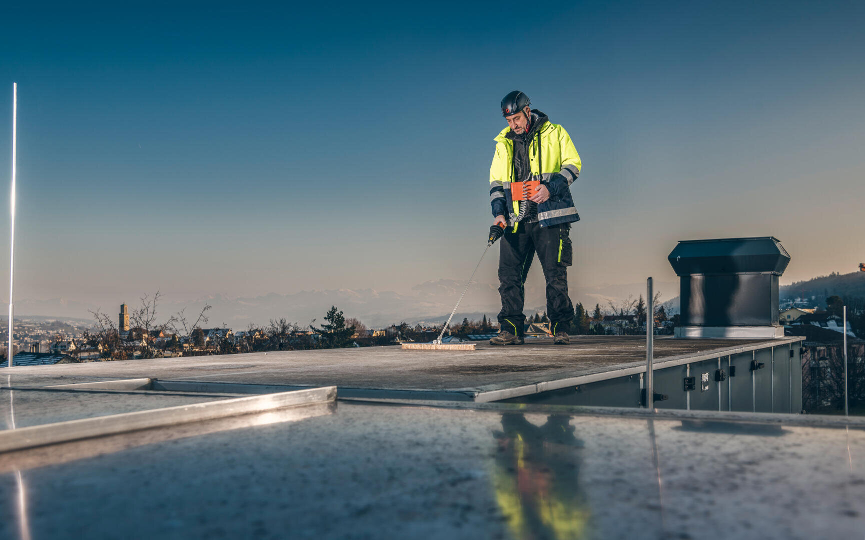 Un ouvrier en gilet de sécurité et casque de protection se tient sur un toit et travaille avec un outil. En arrière-plan, par temps clair, on aperçoit les bâtiments de la ville et les montagnes.