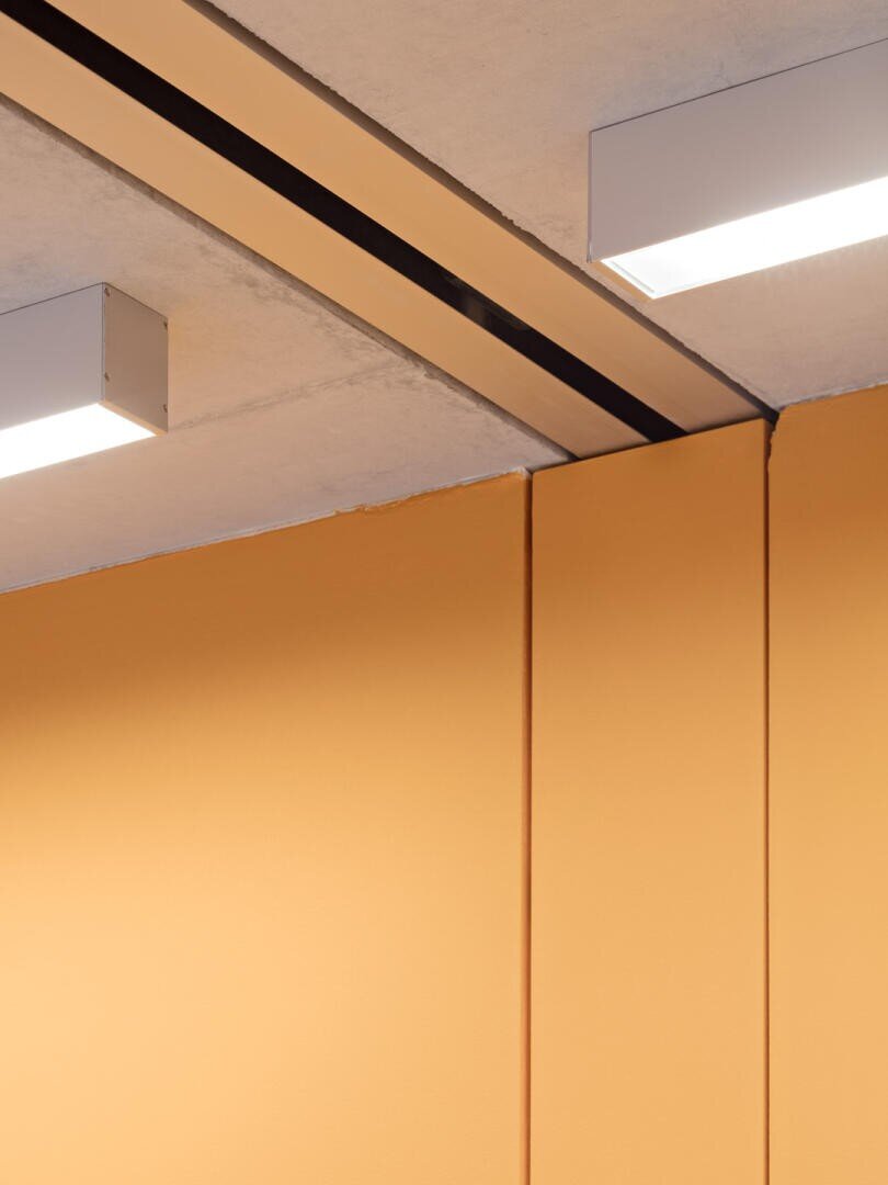 Intérieur minimaliste avec un mur jaune-orange qui rencontre un plafond en béton, avec deux plafonniers rectangulaires et des lignes sombres qui marquent les joints entre les surfaces. Le design est clair et géométrique.