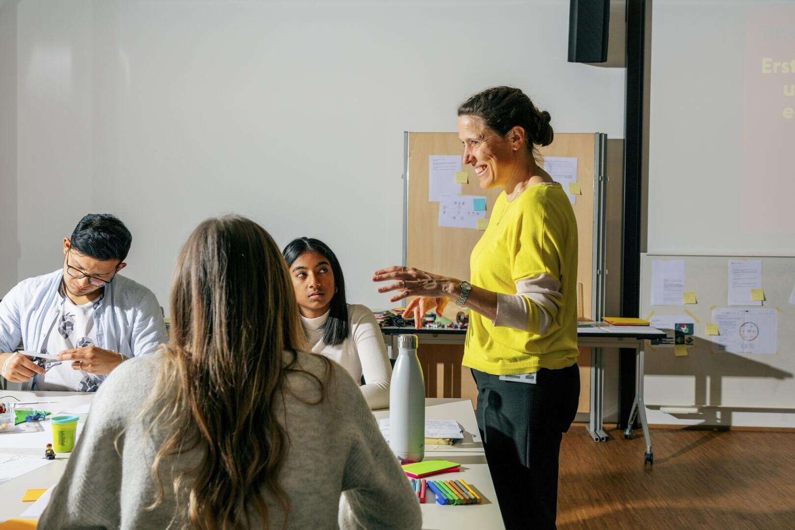 Eine Frau in einem gelben Pullover steht und spricht mit drei Personen, die in einem hell erleuchteten Klassenzimmer um einen Tisch mit Notizbüchern und bunten Stiften sitzen.
