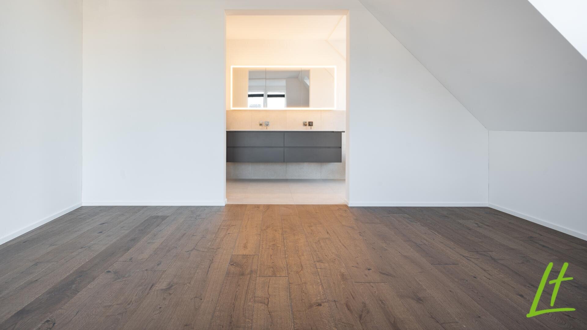 Ein heller, leerer Raum mit dunklem Holzboden und weißen Wänden. Eine offene Tür gibt den Blick auf ein modernes Badezimmer mit einem breiten Spiegel und Unterschränken frei. Der Raum hat eine schräge Decke und ist minimalistisch eingerichtet.