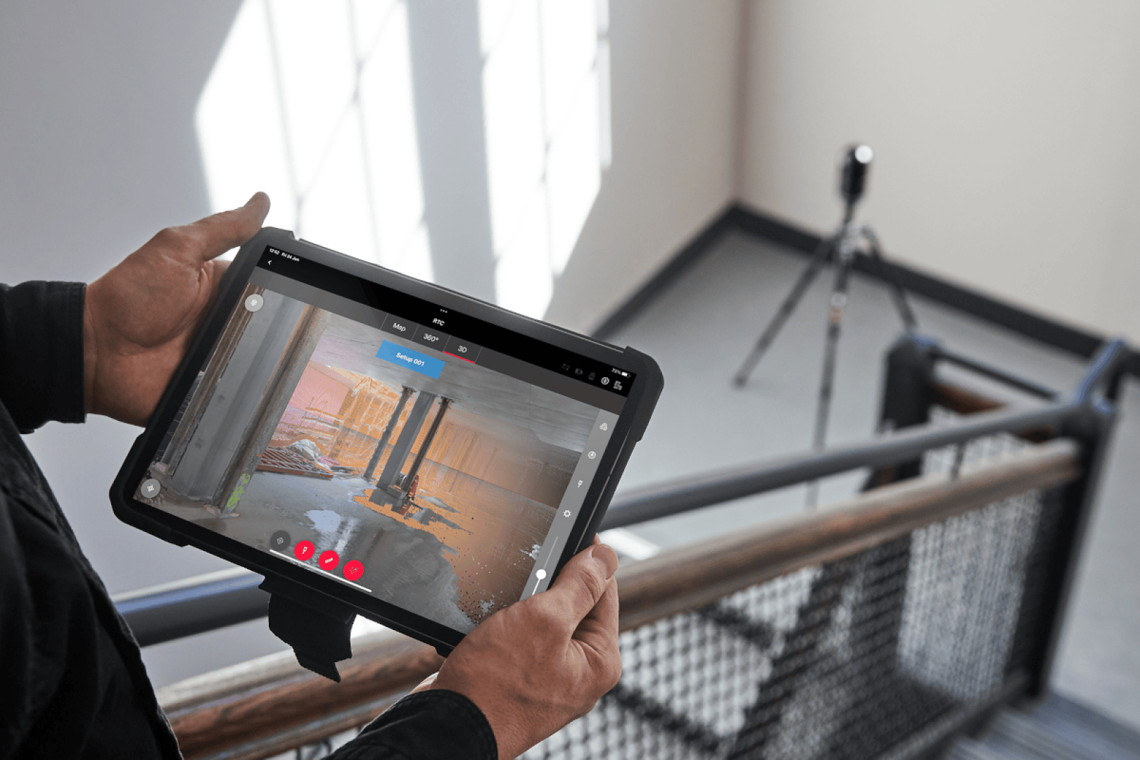 Eine Person hält ein Tablet, das einen 3D-Scan eines Raums mit Säulen zeigt, während ein Laserscanner auf einem Stativ am oberen Ende eines Treppenhauses steht. Sonnenlicht strömt durch die Fenster und beleuchtet die Szene.