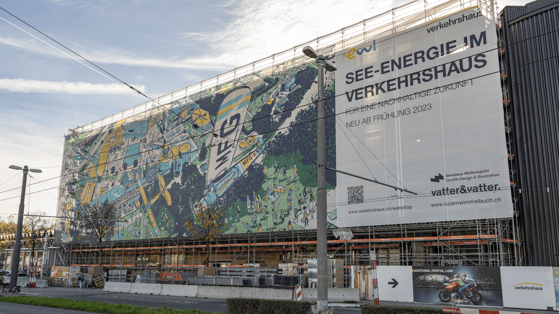 Ein großes, im Bau befindliches Gebäude ist mit Gerüsten und einem farbenfrohen, illustrierten Banner bedeckt, das für die Ausstellung See-Energie im Verkehrshaus wirbt, die im Frühjahr 2023 eröffnet wird, in deutscher Sprache. Davor verlaufen Straßen- und Straßenbahnschienen.
