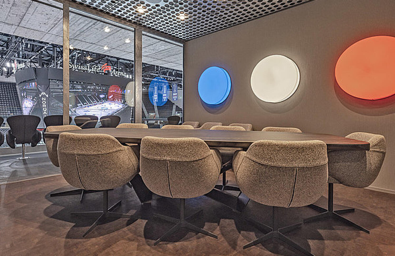Une salle de conférence moderne avec une grande table en bois entourée de chaises rembourrées. Trois luminaires circulaires bleus, blancs et rouges décorent le mur, et de grandes fenêtres donnent sur un stade couvert.