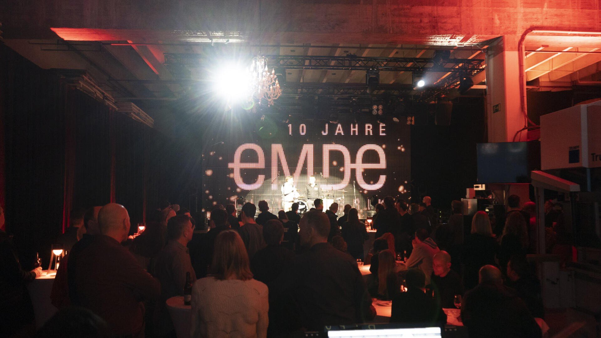 Eine Menschenmenge nimmt an einer Indoor-Veranstaltung mit schummriger Beleuchtung teil und steht vor einer großen Bühne, auf der der Schriftzug "10 Jahre embe" leuchtet. Die Atmosphäre ist festlich mit roter Beleuchtung und Kronleuchtern über dem Kopf.