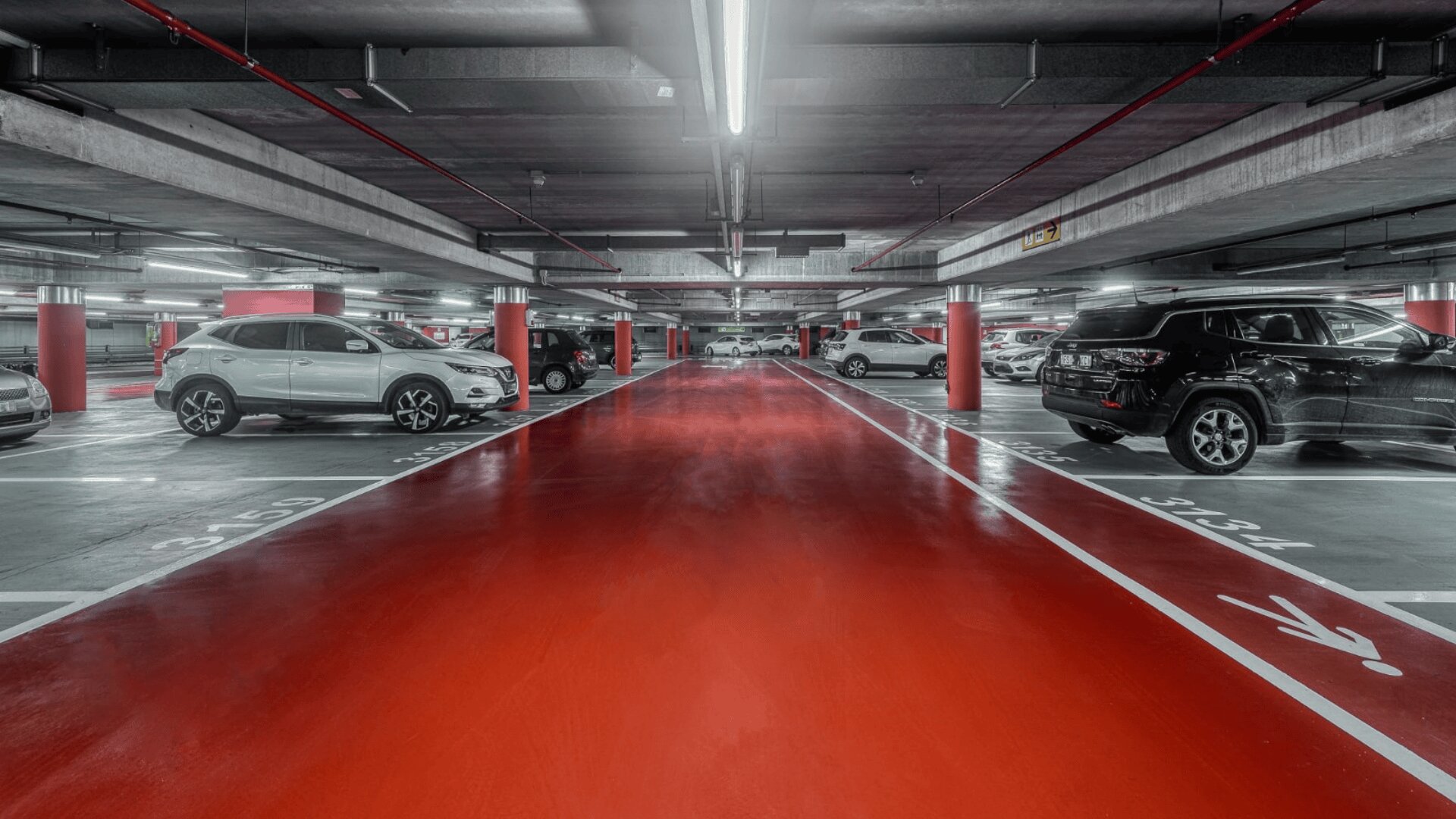 Garage souterrain avec un plafond très éclairé, un sol rouge et plusieurs voitures garées des deux côtés. Les lignes et les numéros des places de stationnement sont visibles en blanc, et les piliers en béton sont peints en rouge.