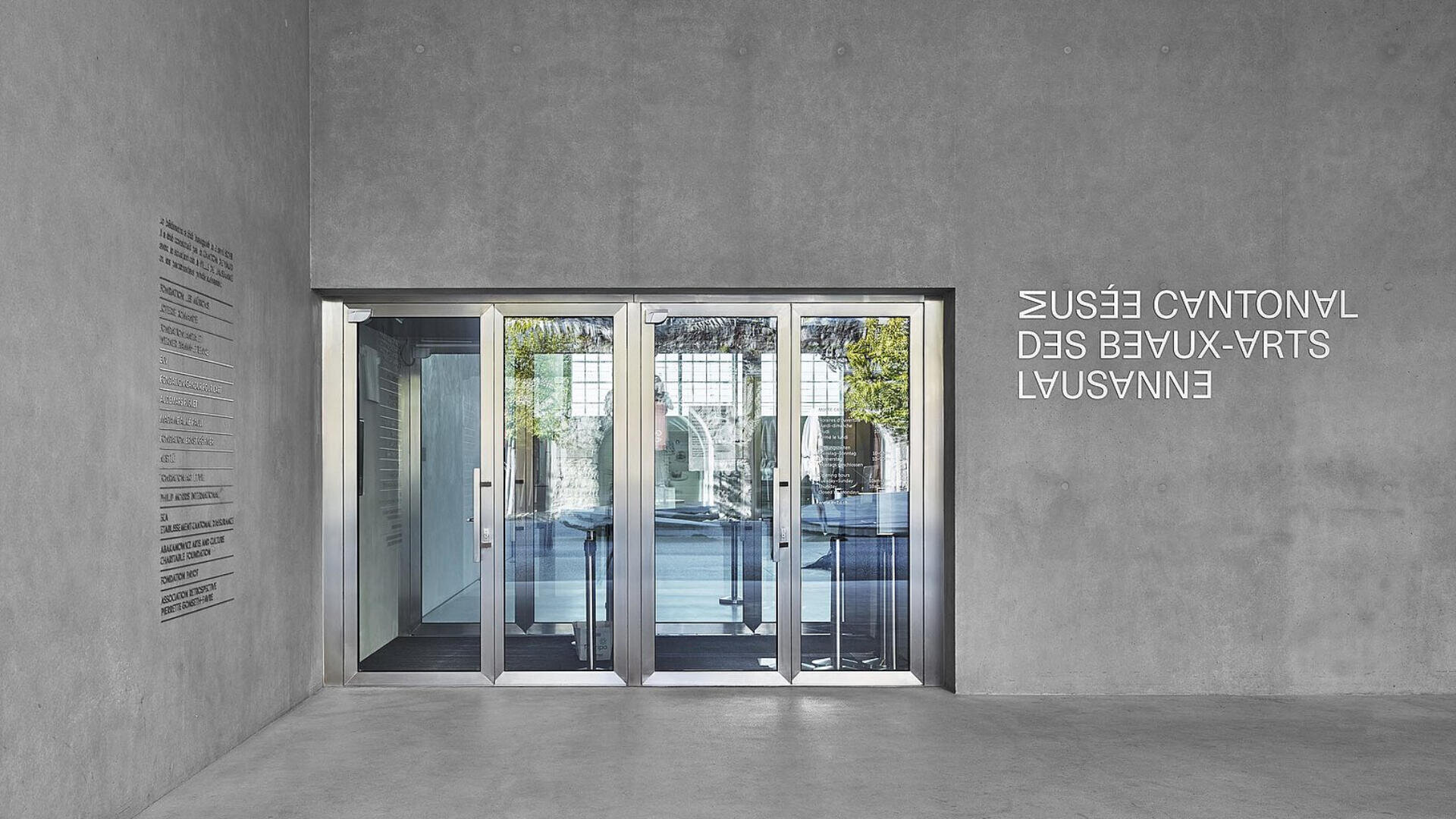Ein moderner Glaseingang mit Schiebetüren in einem Betongebäude; draußen sind Spiegelungen und Grünflächen zu sehen. Der Text an der Wand lautet MUSÉE CANTONAL DES BEAUX-ARTS LAUSANNE in umgekehrter Reihenfolge, und auf der linken Seite befindet sich ein Verzeichnis.