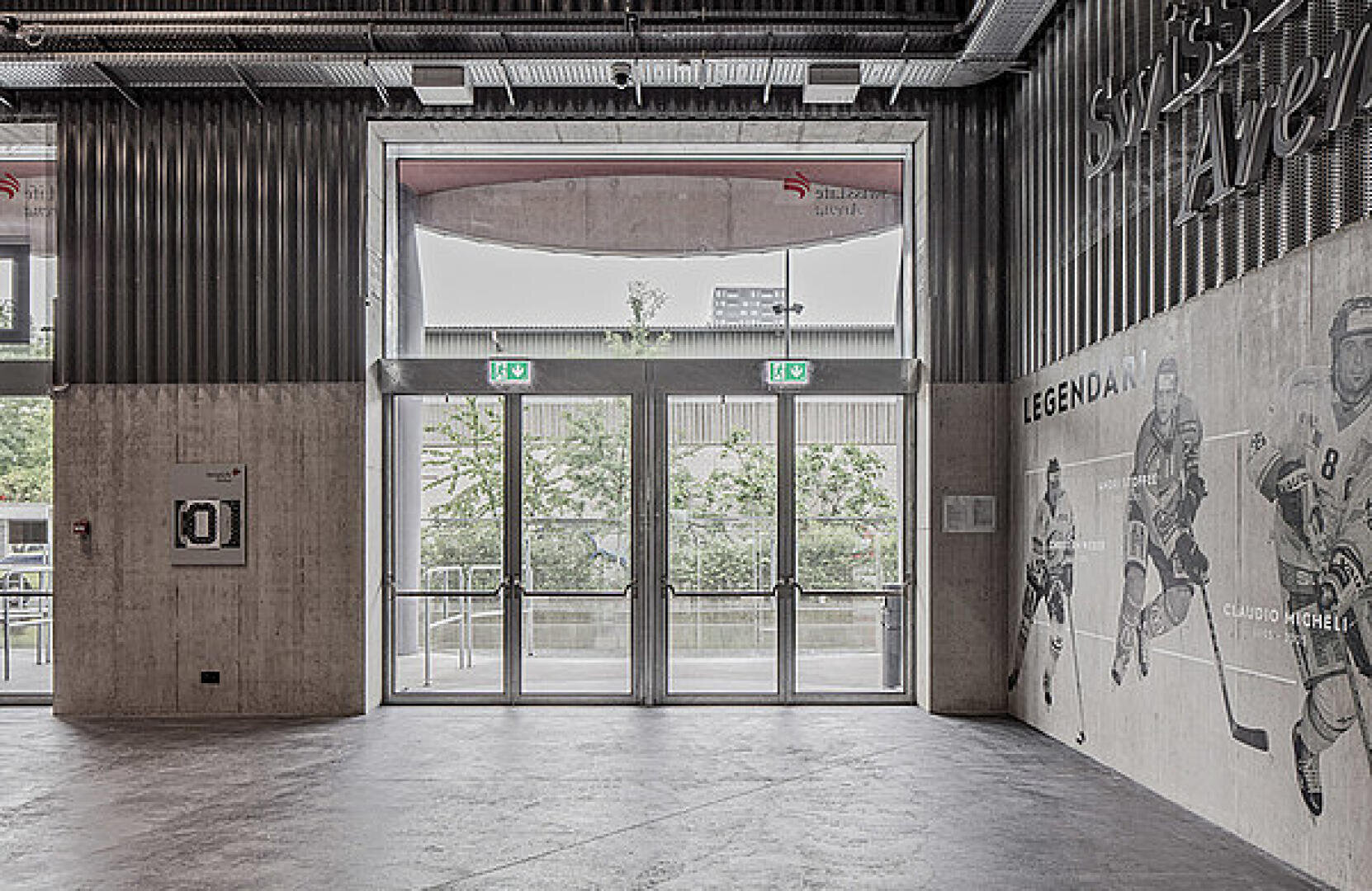 Une entrée de bâtiment moderne avec de grandes portes vitrées, des panneaux de sortie de secours et une fresque de joueurs de hockey sur glace sur le mur de droite ; de la verdure et un bâtiment sont visibles à travers les portes.