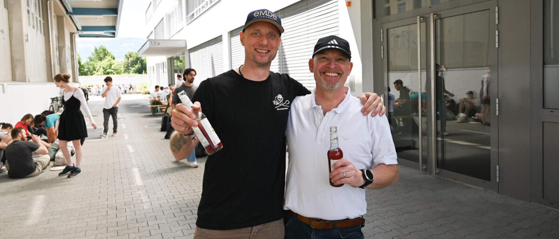 Zwei lächelnde Männer mit Getränken in der Hand stehen Arm in Arm im Freien, umgeben von anderen Menschen, die auf einem schattigen Gehweg in der Nähe eines modernen Gebäudes sitzen oder stehen.
