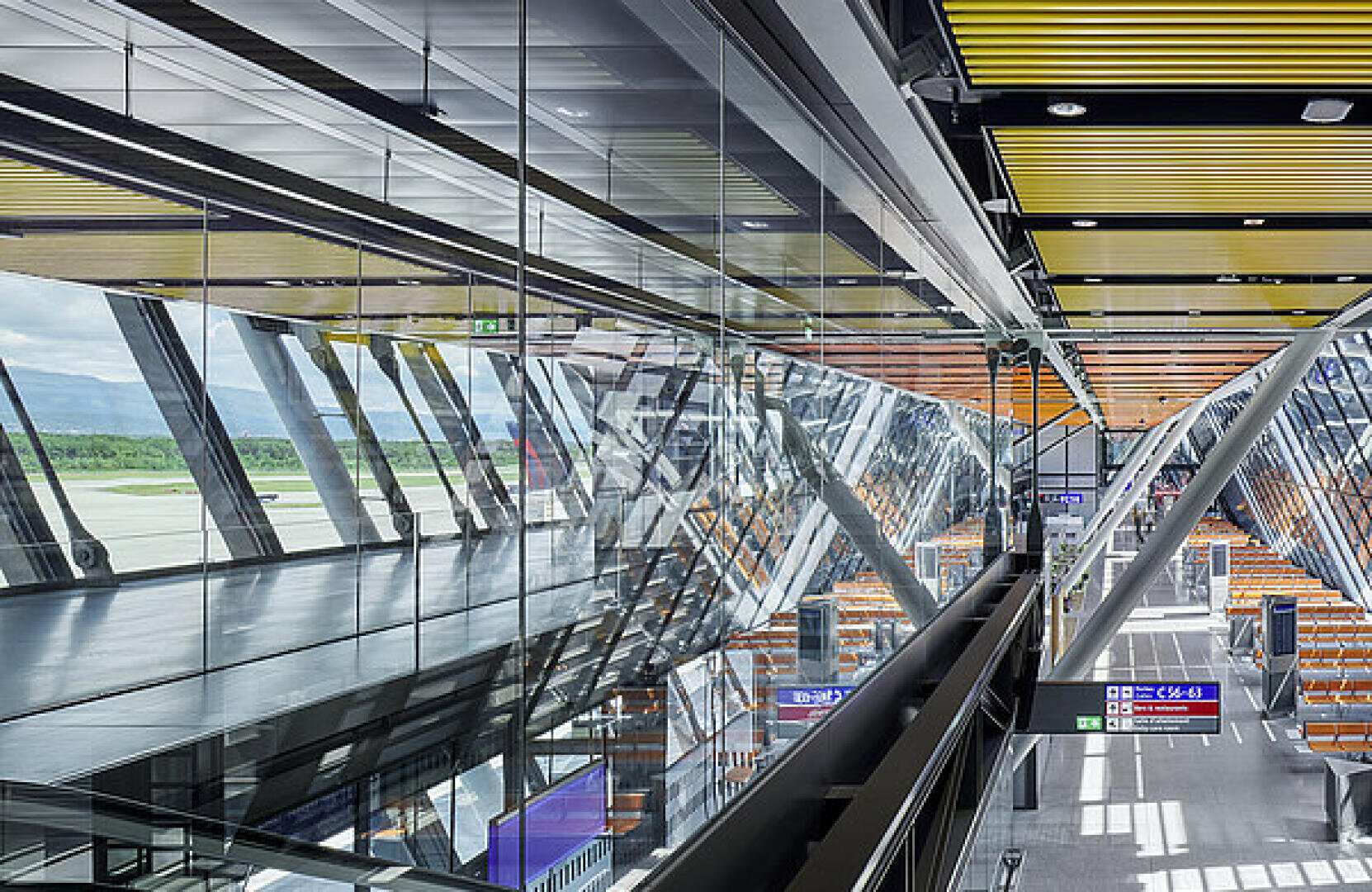 Modernes Flughafenterminal mit Glaswänden, Metallträgern und gelben Deckenakzenten. Man sieht Reihen leerer orangefarbener Sitze und Schilder für Abflüge sowie große Fenster mit Blick auf die Startbahn und die Landschaft draußen.