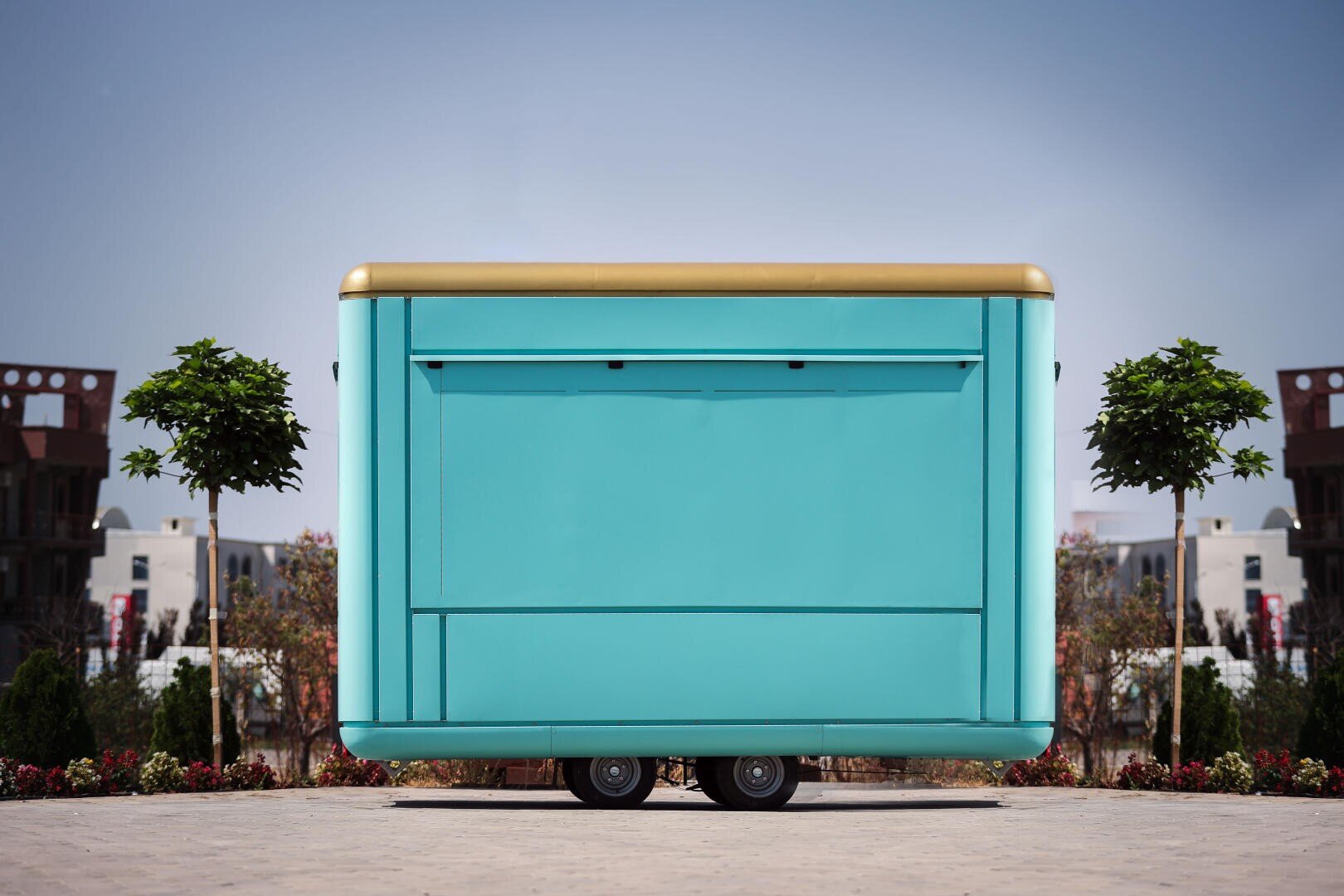 Un camion-restaurant mobile de couleur sarcelle et or, aux fenêtres de service fermées, est garé sur une surface pavée, flanqué de deux petits arbres, avec des bâtiments et des espaces verts en arrière-plan sous un ciel dégagé.