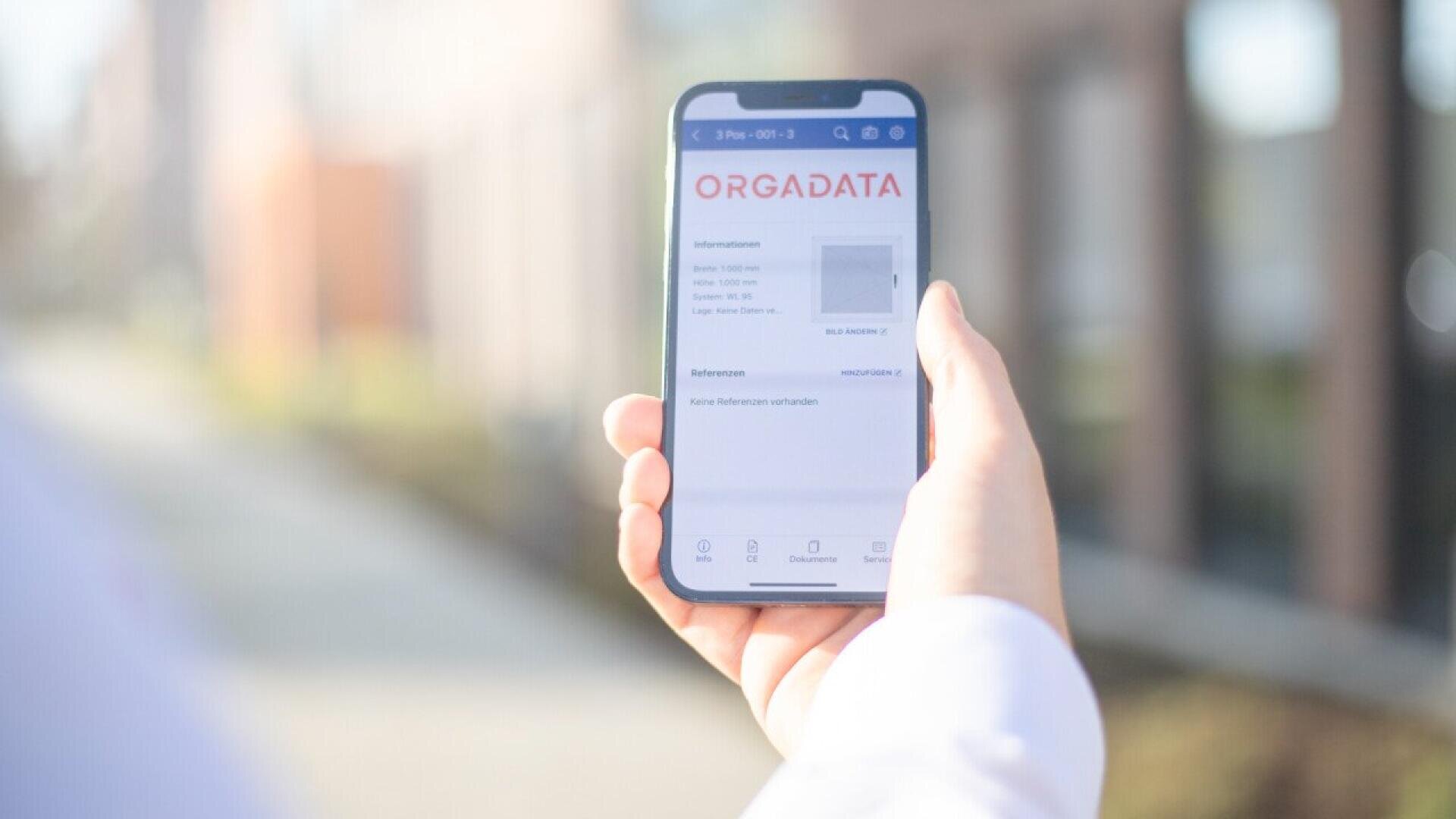 Une personne tenant un smartphone affichant une application avec le titre ORGADATA sur l'écran, en extérieur avec un arrière-plan flou.