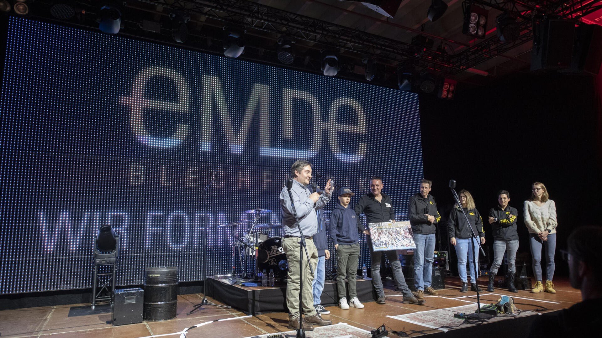 Eine Gruppe von Personen steht auf einer Bühne, hinter der ein großer LED-Bildschirm "emde" und "Wir Fortschritt" anzeigt. Eine Person spricht in ein Mikrofon, während andere in der Nähe stehen, einige halten einen großen gerahmten Gegenstand.