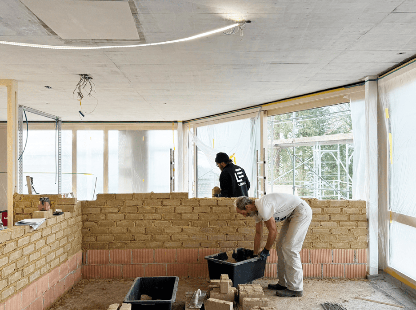 Zwei Arbeiter bauen eine Innenwand aus Ziegelsteinen in einem halbfertigen Raum mit großen Fenstern. Ein Mann beugt sich über einen Eimer, während der andere in der Nähe der Wand steht. Baumaterialien und Werkzeuge liegen verstreut herum.