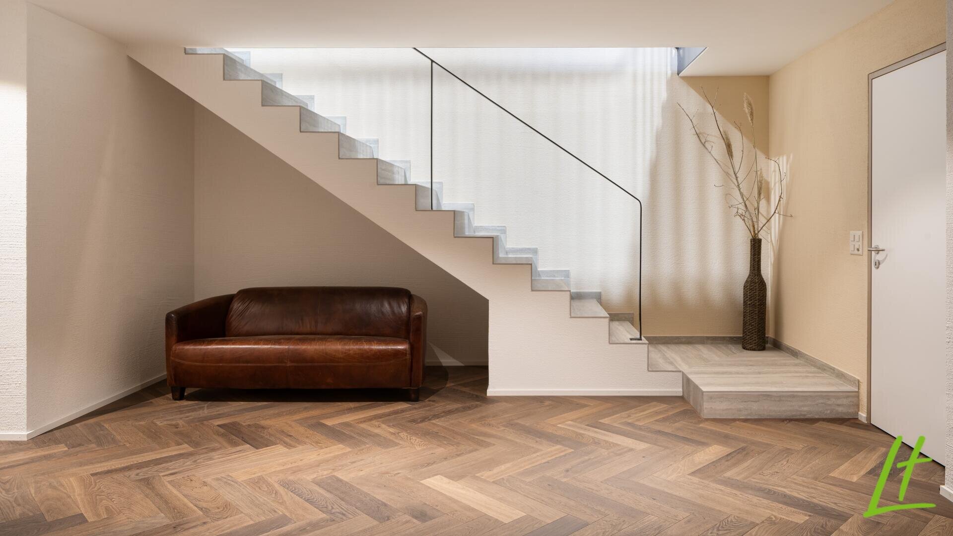 Un intérieur moderne avec un canapé en cuir marron sous un escalier flottant avec une rampe en verre. Le sol est en bois à chevrons et un grand vase avec des branches se trouve près d'une porte blanche.