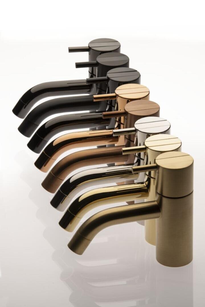 Eine Reihe moderner, zylindrischer Wasserhähne in verschiedenen Metallic-Farben, darunter Schwarz, Bronze, Kupfer, Chrom und Gold, die diagonal auf einer reflektierenden weißen Oberfläche angeordnet sind.