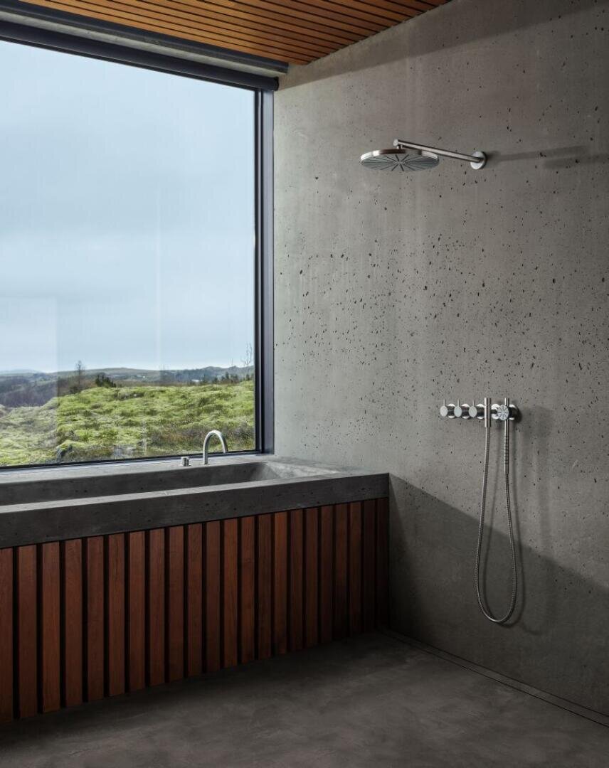 Ein modernes Badezimmer mit einer Betonwand, minimalen Duscharmaturen, einer holzverkleideten Badewanne und einem großen Fenster mit Blick auf eine Graslandschaft unter einem bewölkten Himmel.