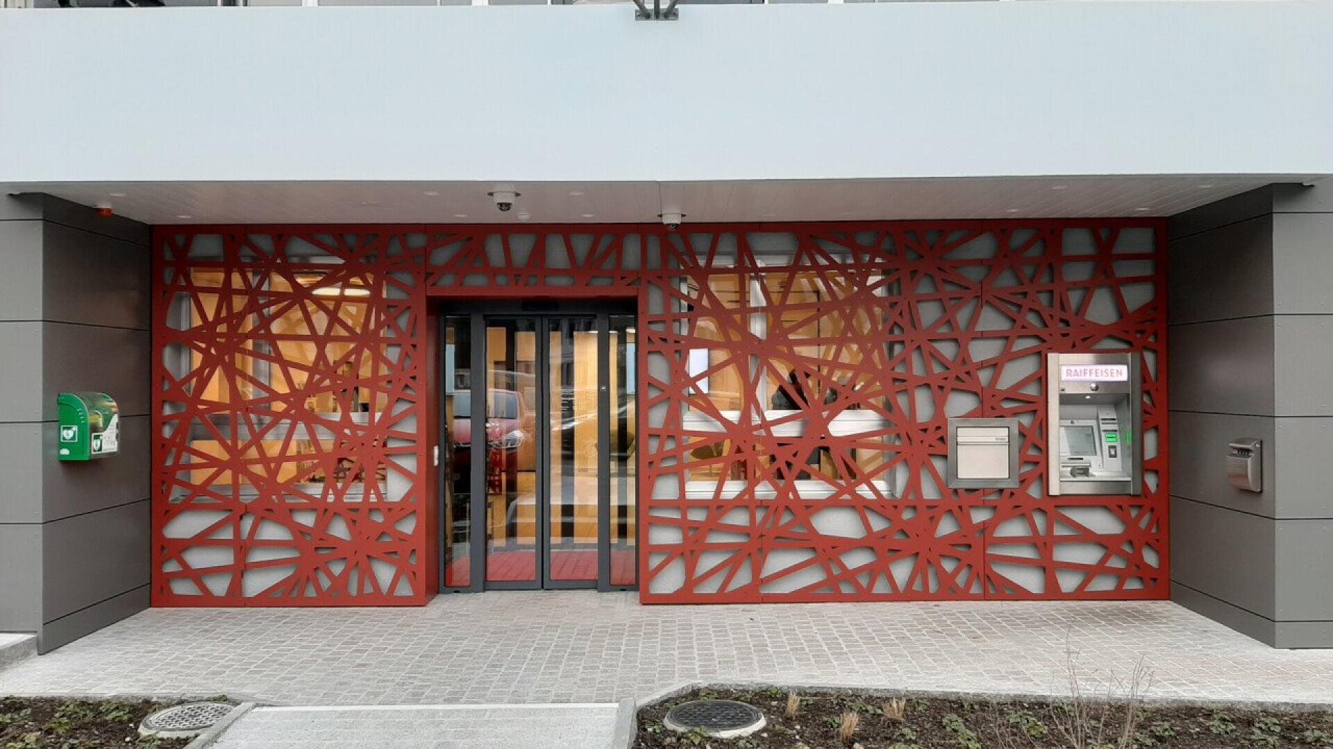 L'entrée d'un bâtiment moderne présente une grille métallique rouge décorative avec un motif abstrait, des portes vitrées au centre, un distributeur automatique de billets et des boîtes aux lettres à droite, et un défibrillateur à gauche.