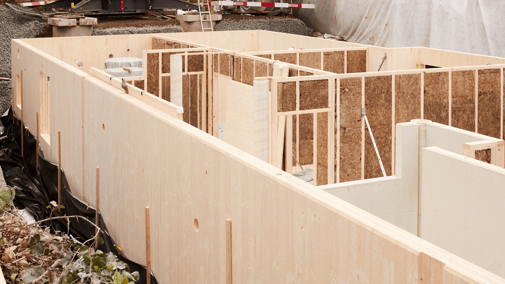 Teilweise errichtetes Holzhausfundament mit freigelegtem Dachstuhl und Innenwänden, umgeben von Erde und Baumaterialien auf einer Baustelle.