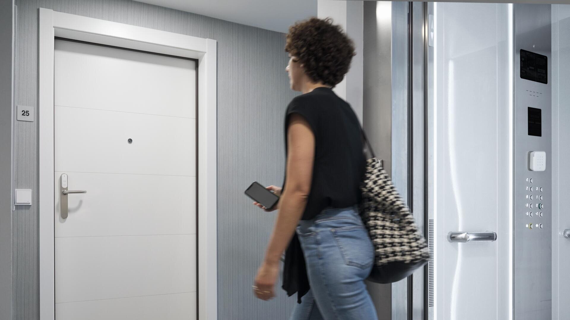 Eine Person mit lockigem Haar, die ein schwarzes Hemd und blaue Jeans trägt, ein Telefon in der Hand hält und eine gemusterte Tasche mit sich führt, geht an einer weißen Tür und einem offenen Aufzug in einem modernen Flur vorbei.
