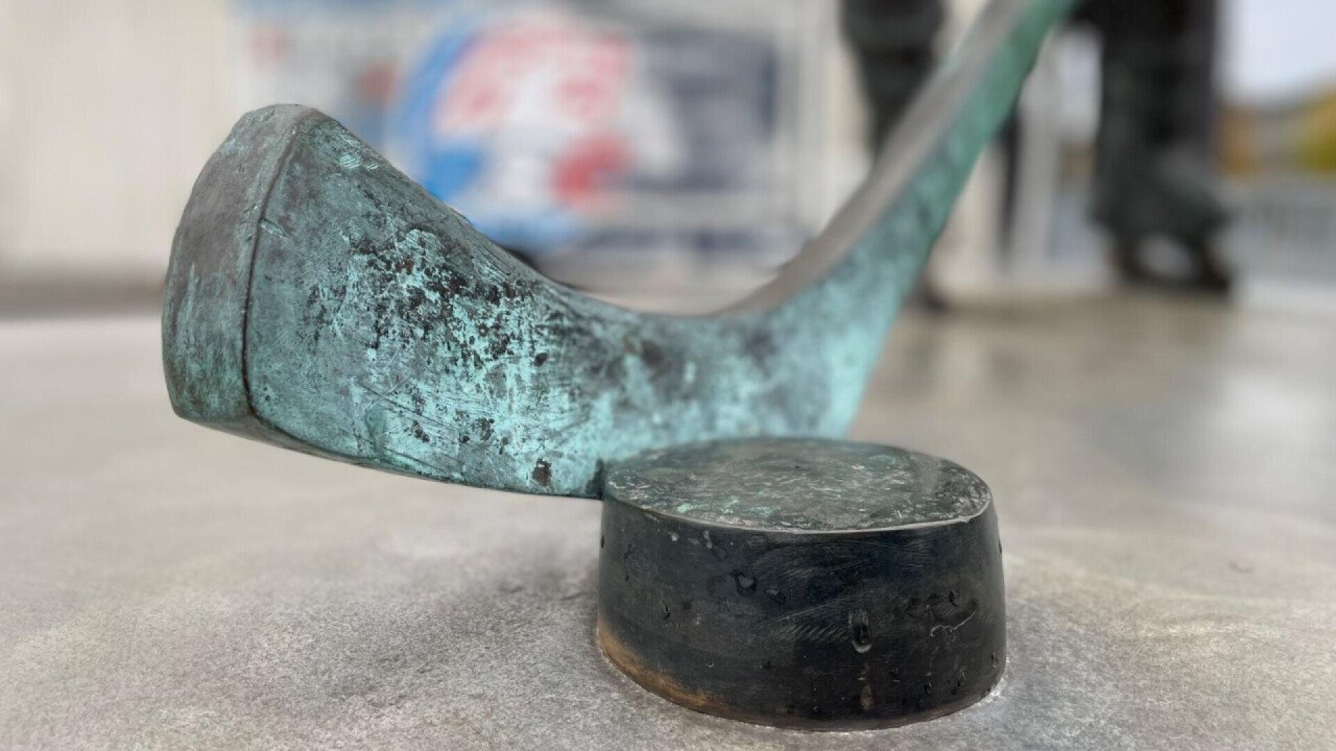 Gros plan d'une crosse de hockey en bronze usée par les intempéries, posée juste au-dessus d'un palet de hockey en bronze sur une surface lisse, avec un arrière-plan flou.