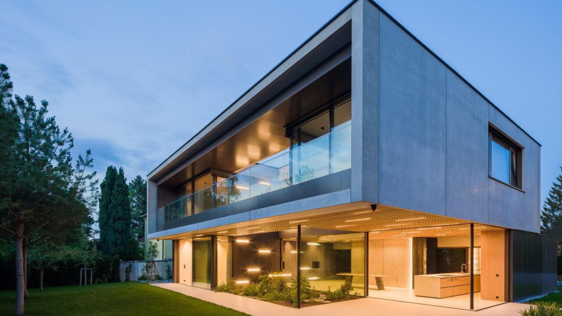 Une maison moderne en béton avec de grandes parois vitrées au niveau inférieur et un balcon avec des garde-corps en verre au niveau supérieur, entourée d'une pelouse verte et d'arbres.
