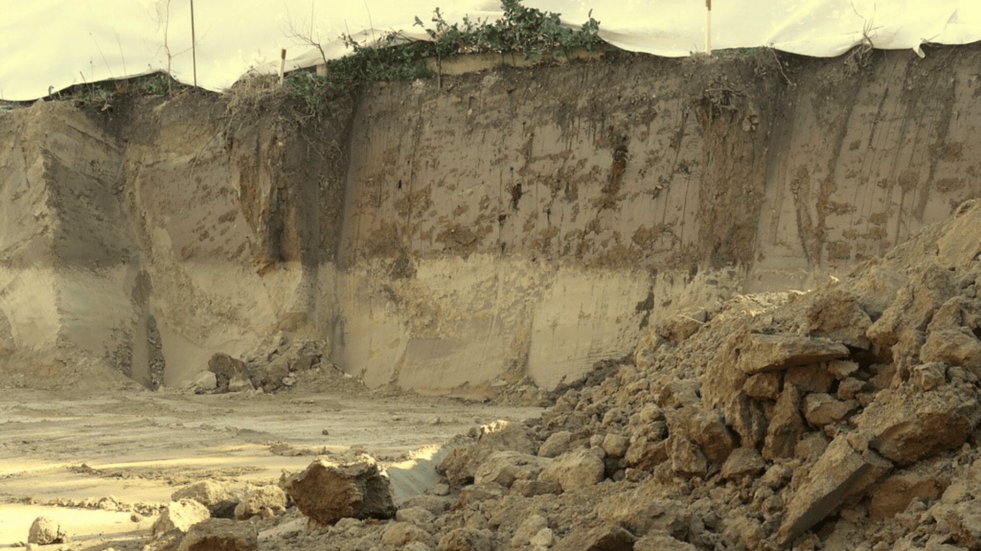 Die Nahaufnahme einer Baustelle zeigt eine freiliegende Erd- und Lehmwand mit sichtbaren Schichten und verstreutem losem Boden und Steinen an der Basis. Am oberen Rand wächst eine spärliche Vegetation.