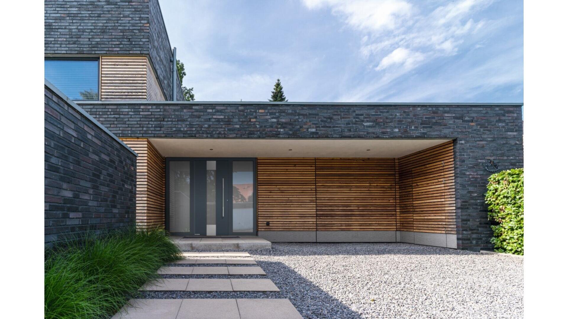 Extérieur d'une maison moderne avec des briques foncées et des lattes de bois horizontales, une grande porte d'entrée en verre dépoli, une allée pavée, une zone de gravier et des buissons verts sous un ciel partiellement nuageux.