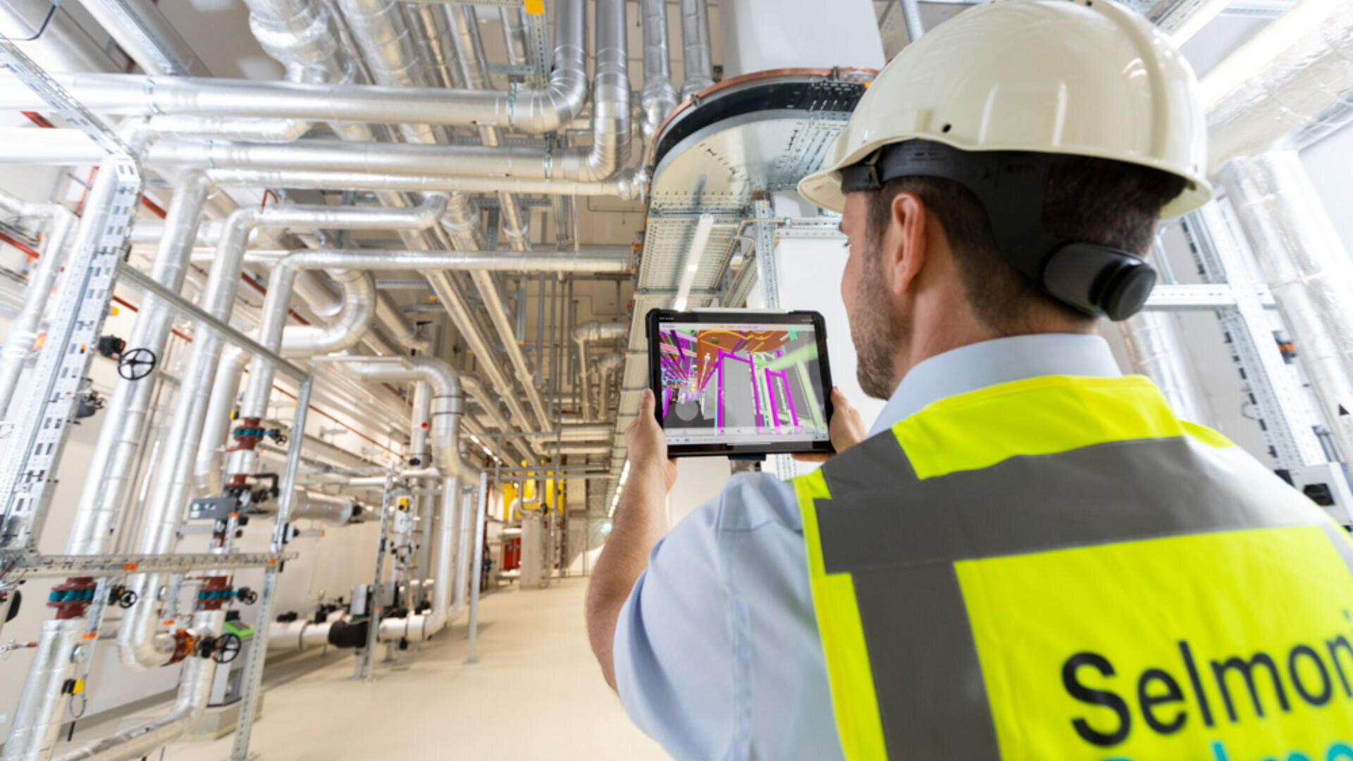 Ein Bauarbeiter, der einen Schutzhelm und eine Warnweste trägt, verwendet ein Tablet, um ein digitales Modell zwischen komplexen Rohren und Kanälen in einer Industrieanlage zu betrachten.