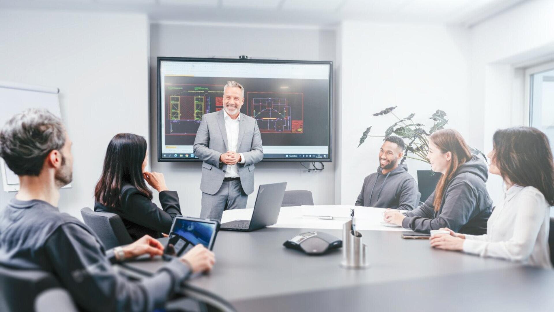 Dans un bureau moderne, un homme en costume se lève et fait une présentation à quatre collègues assis autour d'une table de conférence, avec des diagrammes architecturaux affichés sur un grand écran derrière lui.