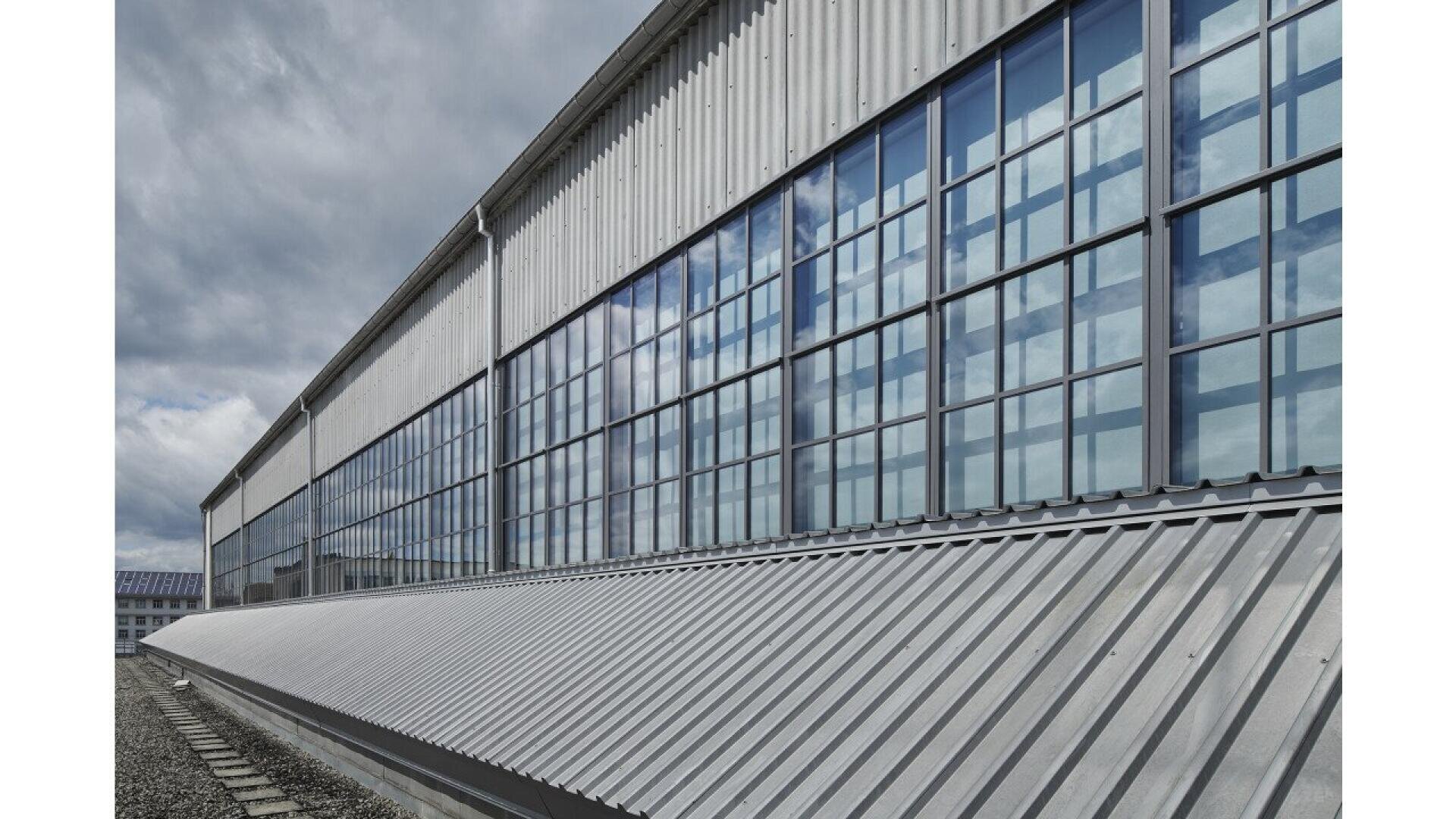 Un grand bâtiment industriel avec de hautes fenêtres à cadre métallique reflétant un ciel nuageux et un toit en tôle ondulée longeant la façade.
