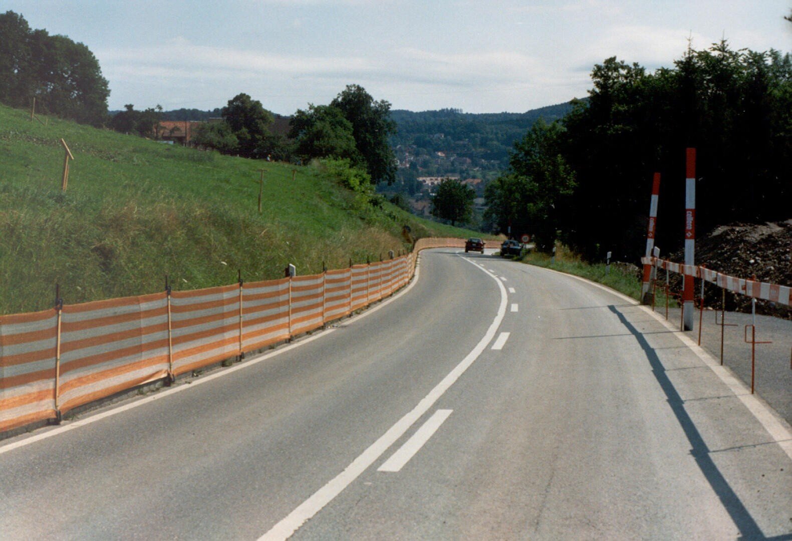 Une route de campagne sinueuse avec des barrières de chantier orange et blanches d'un côté, bordée d'herbe et d'arbres, avec quelques voitures au loin sous un ciel partiellement nuageux.