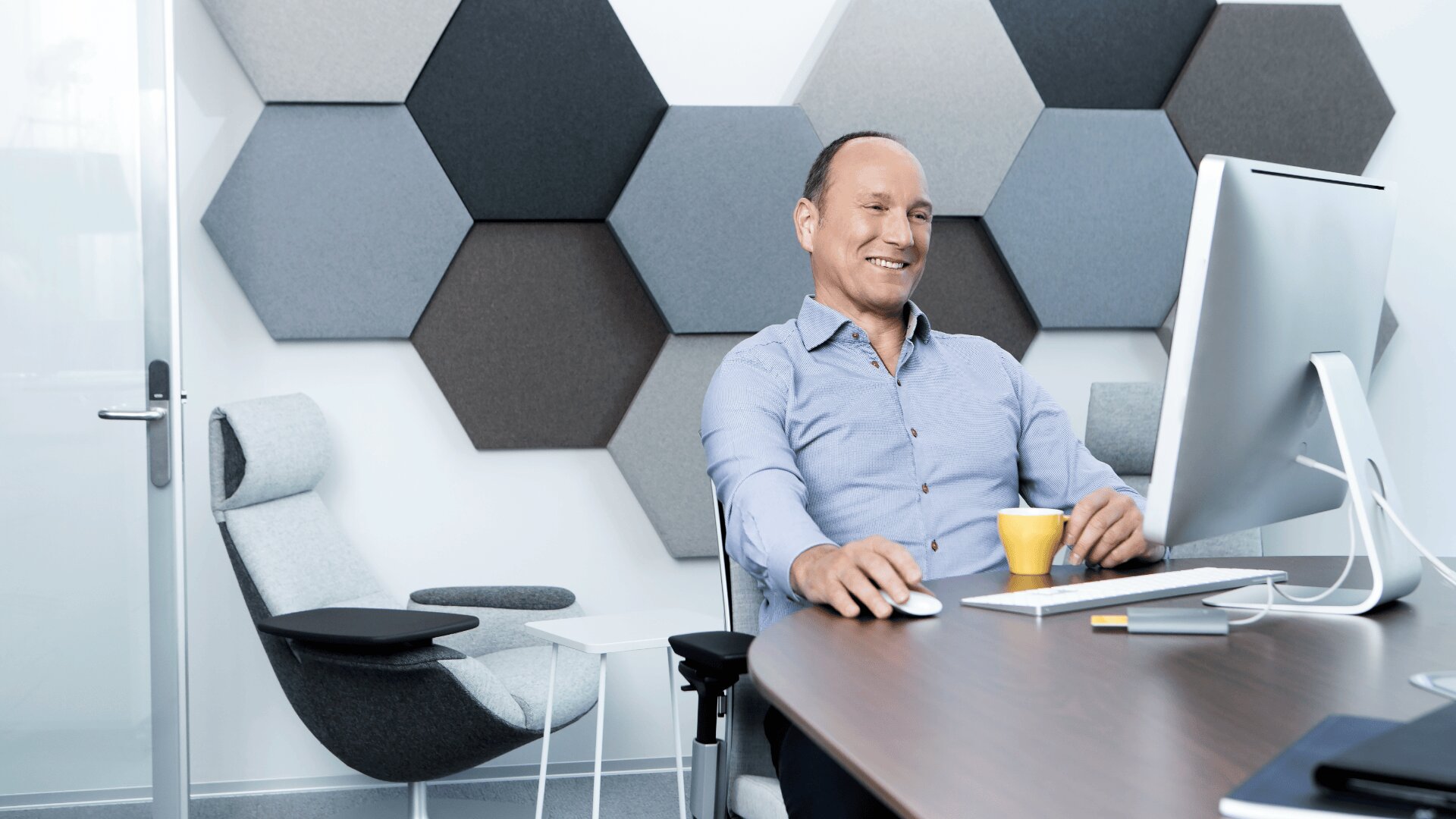 Ein Mann in einem blauen Hemd sitzt an einem Schreibtisch mit einem Computer, lächelt und hält eine gelbe Kaffeetasse in der Hand. Das Büro ist modern eingerichtet, mit sechseckigen Wandpaneelen und modernen Stühlen.