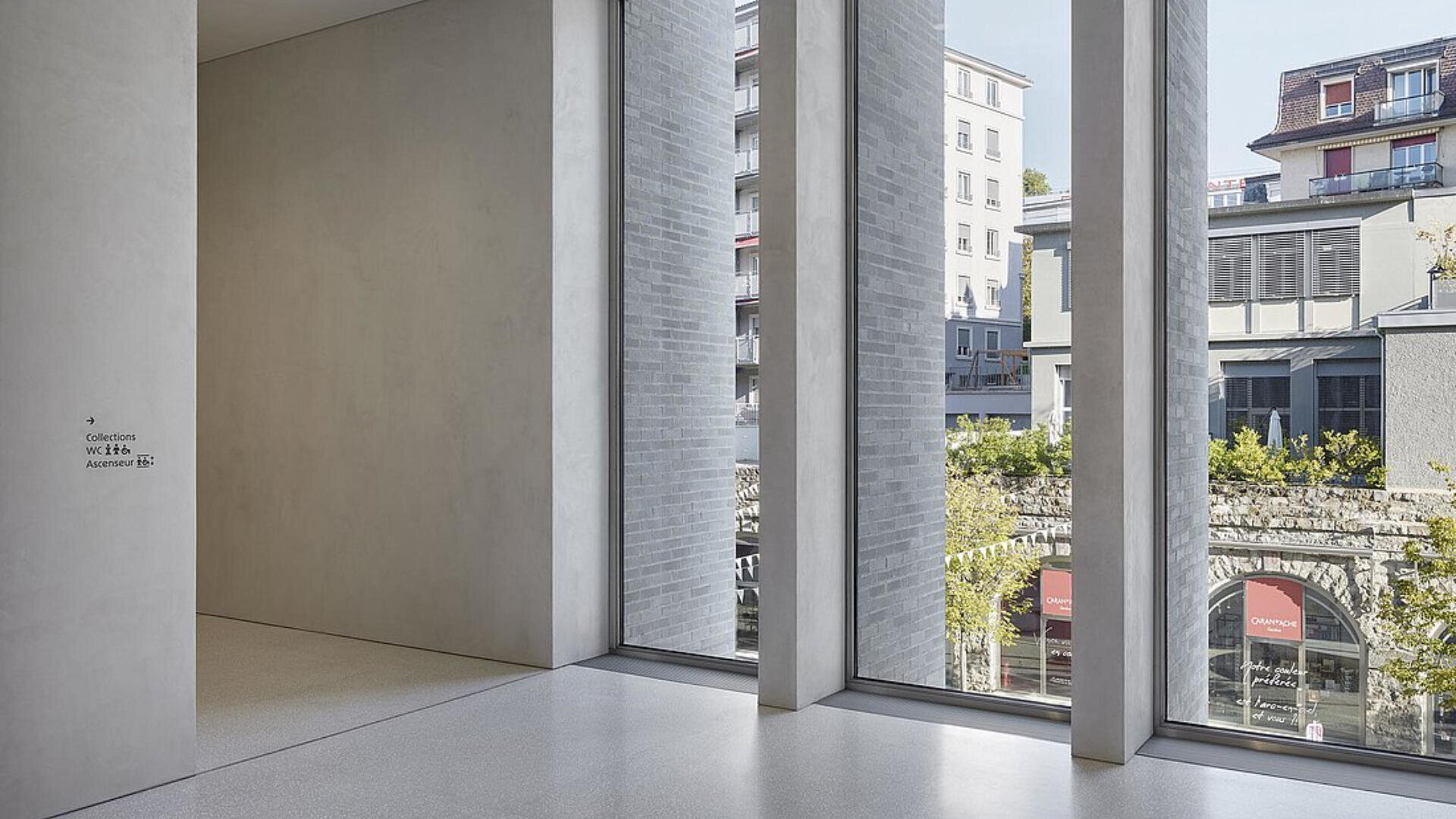 Ein modernes, minimalistisches Interieur mit hohen Fenstern, die den Blick auf die Gebäude der Stadt und die Grünanlagen draußen freigeben. Sanftes natürliches Licht erfüllt den Raum und hebt glatte graue Wände und einen hellen Boden hervor. Schilder weisen den Weg zu den Sammlungen und einem Auditorium.