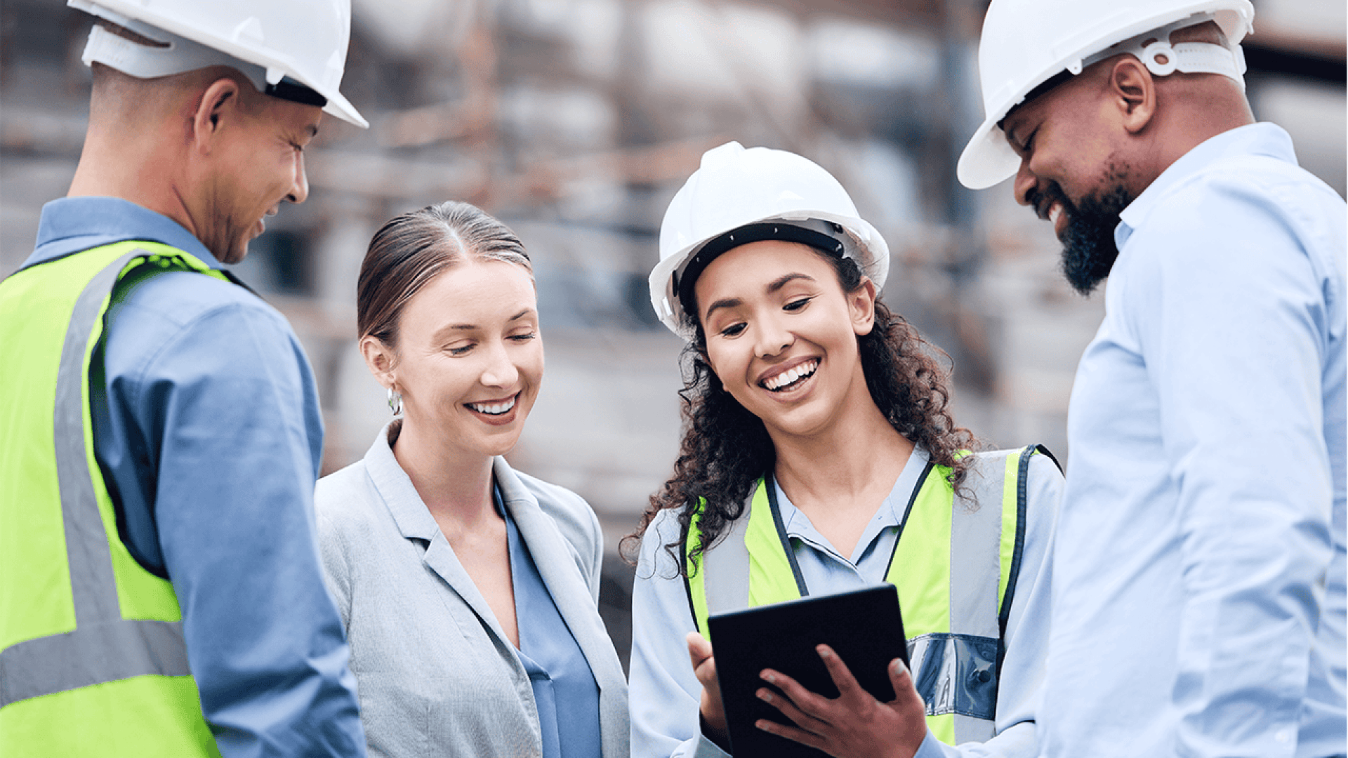 Quatre personnes, deux en gilet de sécurité et casque de chantier, et deux en tenue de travail, se tiennent ensemble sur un chantier de construction, souriant et regardant une tablette.