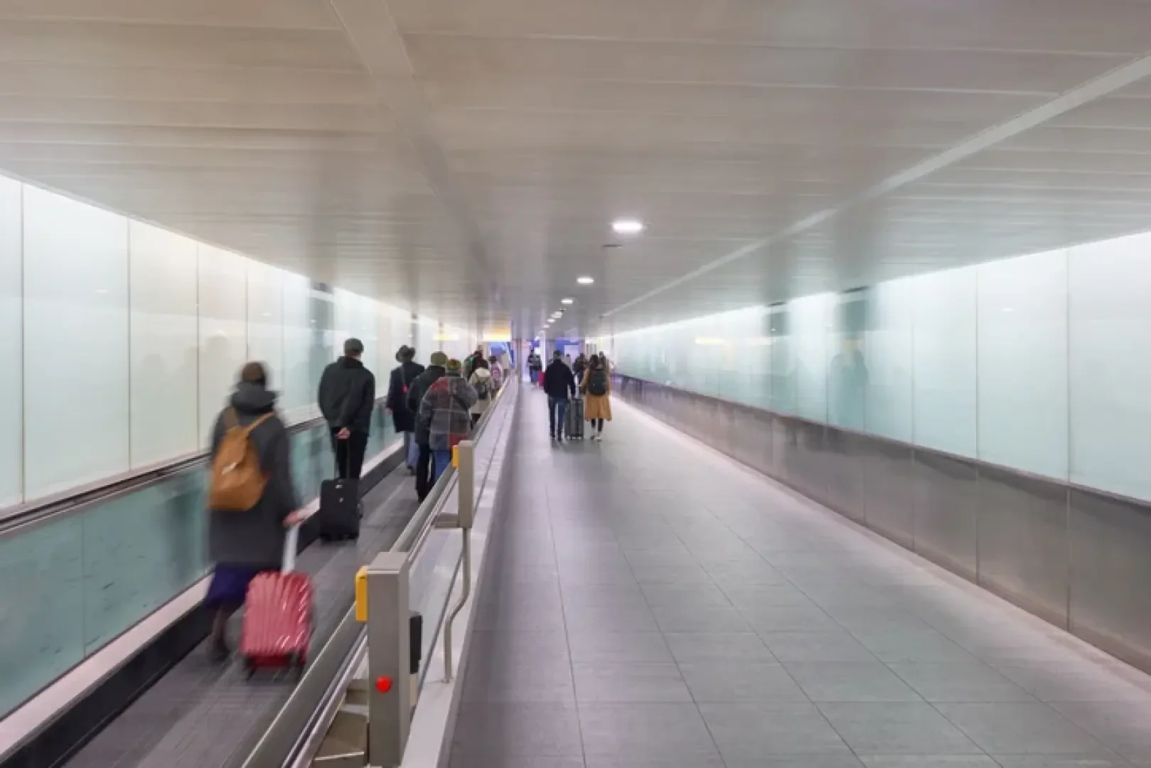 Menschen laufen durch einen langen, hell erleuchteten Flughafenkorridor. Einige benutzen einen Fahrsteig auf der linken Seite, um ihr Gepäck zu tragen, während andere auf der rechten Seite gehen. Der Raum hat Glaswände und einen gefliesten Boden.