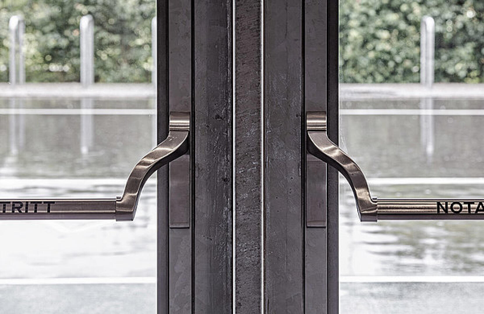 Deux portes vitrées avec poignées argentées, photographiées de l'intérieur. À l'extérieur, une surface humide et une végétation floue suggèrent une pluie récente. Une partie des mots « NOTAU » et « RITT » est visible sur les poignées.