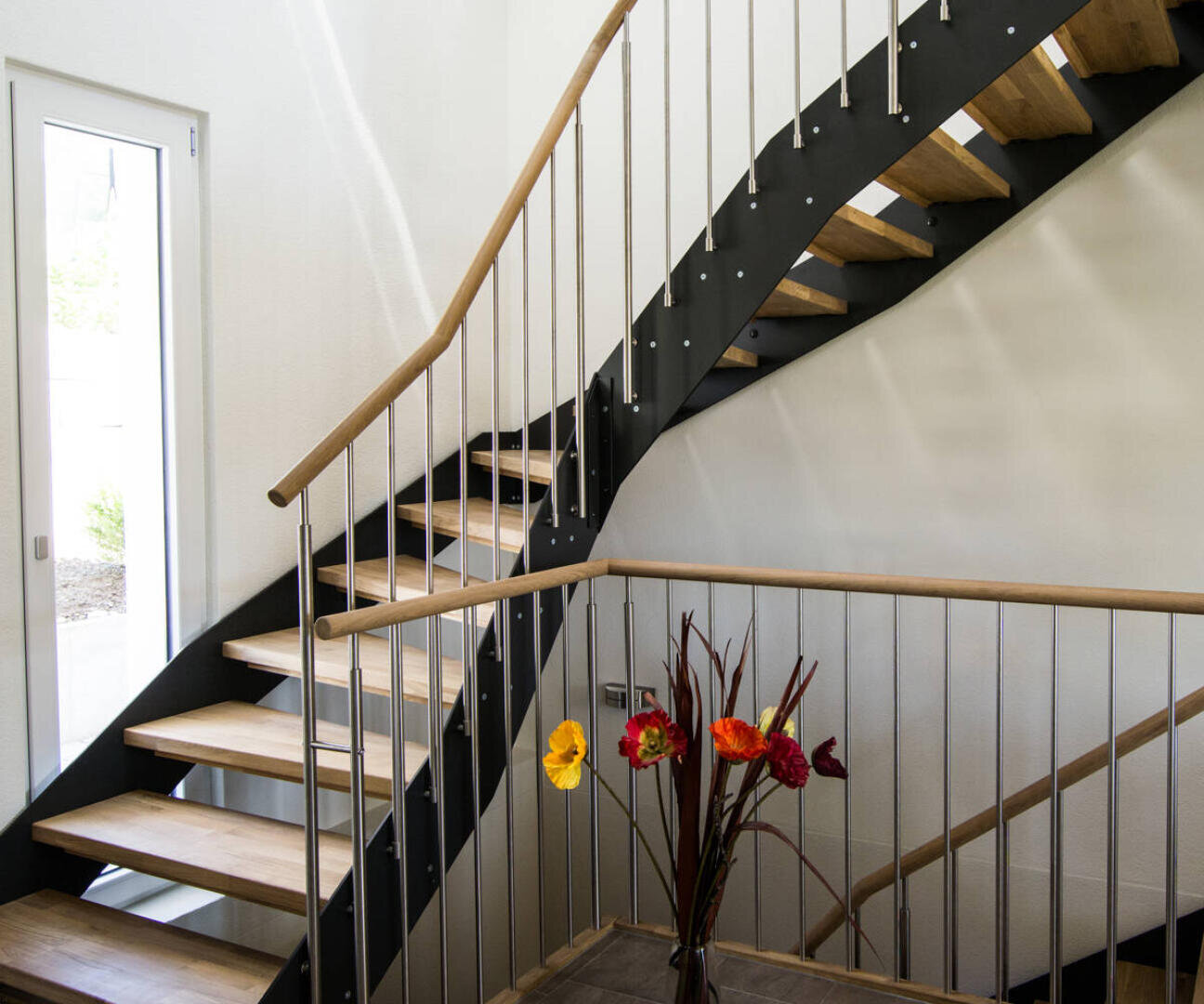 Moderne Innentreppe mit Holzstufen, schwarzen Metallwangen und Metallgeländern. Eine Vase mit bunten Kunstblumen steht auf einem Tisch am unteren Ende der Treppe. Natürliches Licht fällt durch eine Tür auf der linken Seite ein.