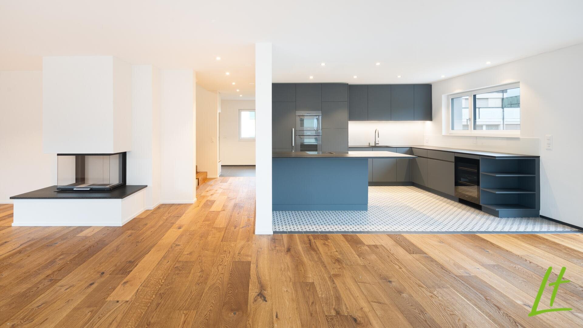 Moderne Küche mit dunkelgrauen Schränken und weißen Arbeitsplatten, offen zum geräumigen Wohnbereich mit Holzböden, einem zeitgenössischen Kamin und viel natürlichem Licht durch große Fenster.