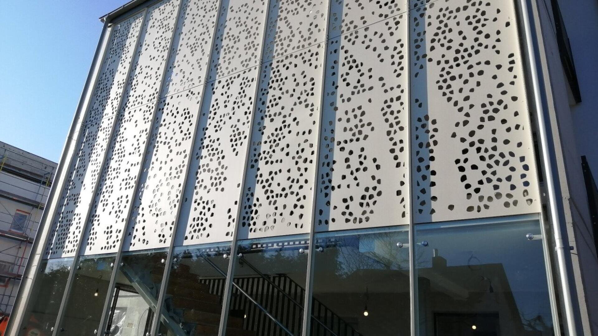 Façade d'un bâtiment moderne avec de hautes fenêtres en verre et des panneaux métalliques décoratifs présentant des motifs de découpe irréguliers. Les panneaux créent un motif en pointillé qui laisse filtrer la lumière. Un escalier est visible derrière les vitres.