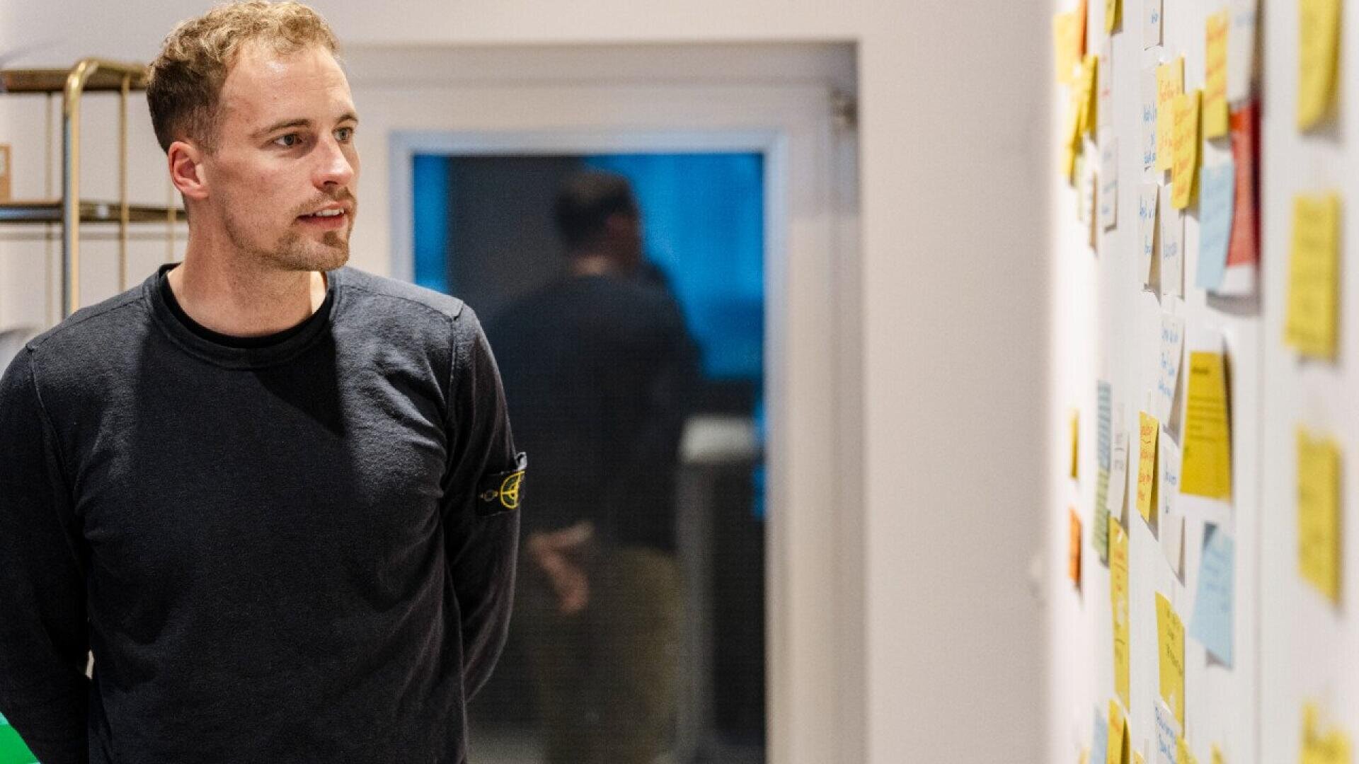 Un homme vêtu d'une chemise noire à manches longues se tient à l'intérieur, regardant un mur couvert de notes adhésives et de papiers colorés. Une fenêtre avec des reflets se trouve à l'arrière-plan.