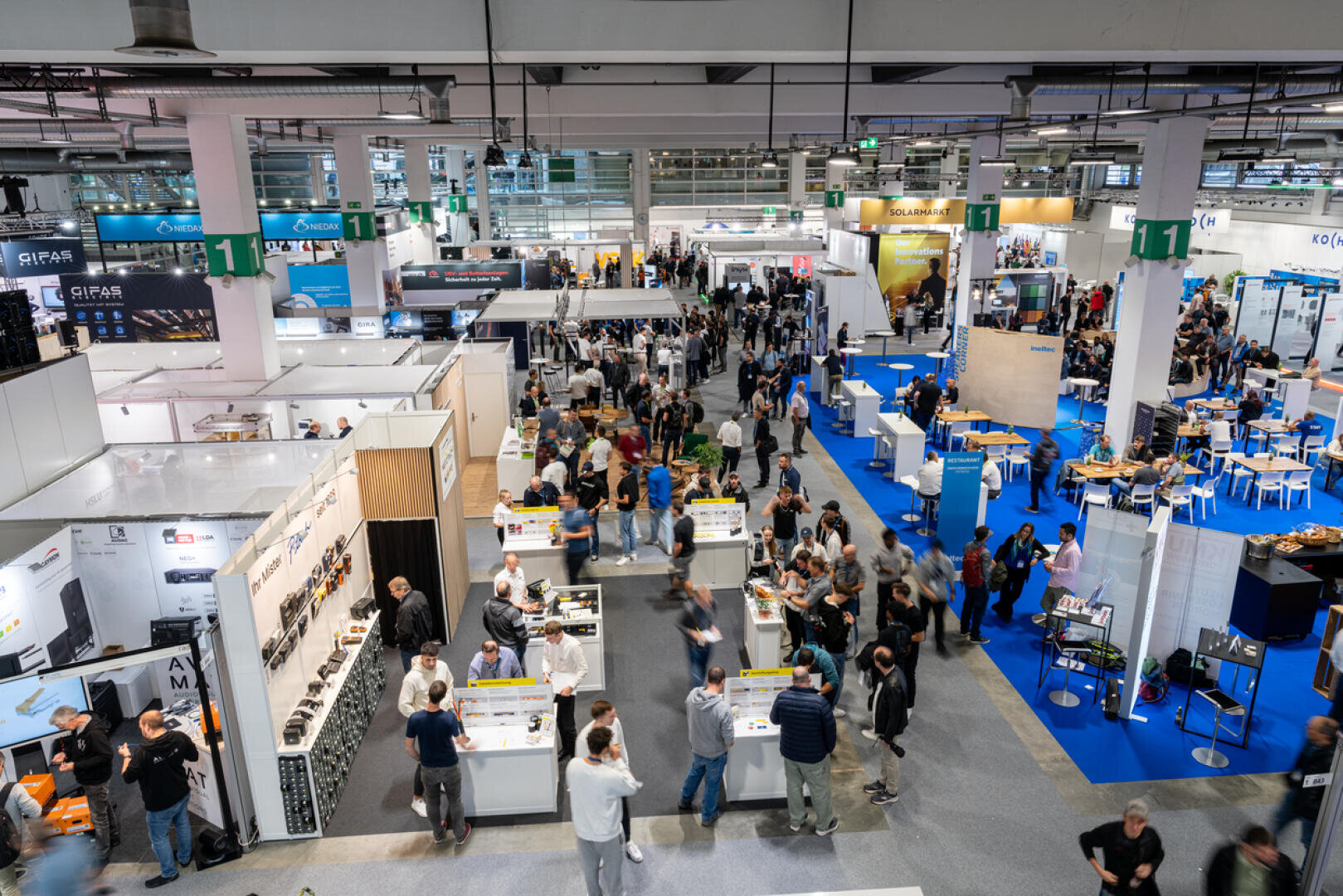 Eine belebte Messe- oder Ausstellungshalle mit vielen Ständen, Ausstellungsstücken und Menschen, die umhergehen, sich unterhalten und die Exponate erkunden. Die Szene spielt in einem Innenraum mit heller Beleuchtung, in dem verschiedene Produkte präsentiert werden.