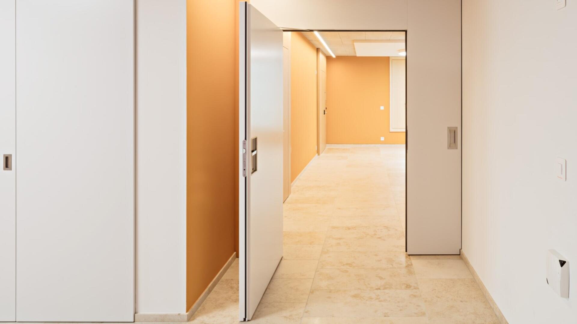 Un couloir moderne et minimaliste avec un sol carrelé beige, des murs d'accent orange et des portes coulissantes blanches, dont l'une est partiellement ouverte et laisse apparaître un espace bien éclairé derrière elle.