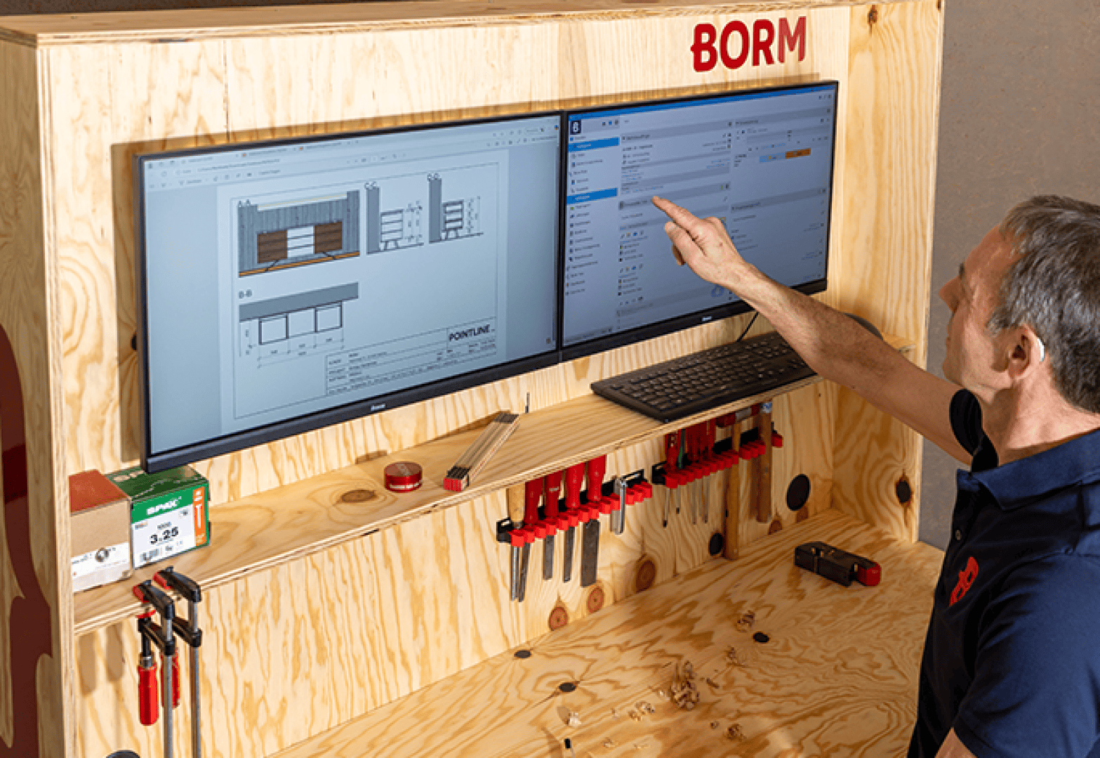 Un homme montre un écran d'ordinateur fixé au mur, sur lequel s'affiche un logiciel de gestion de projet, à côté d'un autre écran sur lequel on peut voir des dessins d'architecture, et en dessous, un poste de travail en bois avec des outils et des pinces.