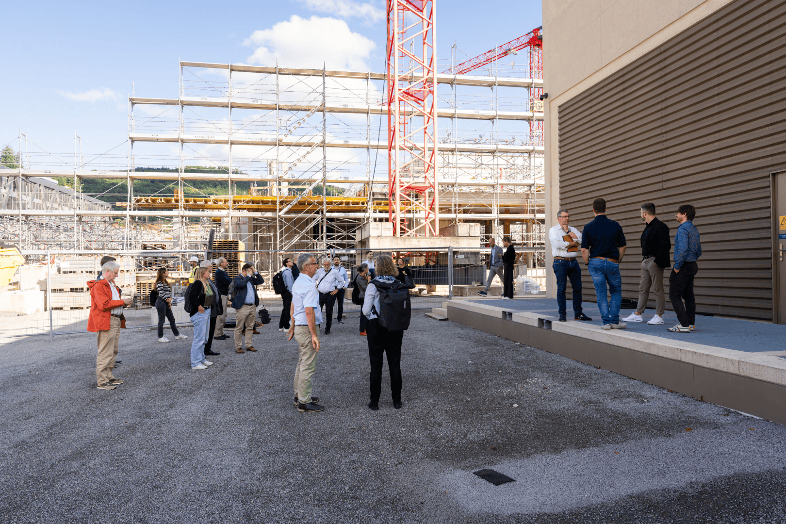 Eine Gruppe von Menschen steht und unterhält sich in der Nähe eines Gebäudes auf einer Baustelle. Im Hintergrund sind Gerüste, Kräne und unfertige Strukturen unter einem blauen Himmel mit Wolken zu sehen.