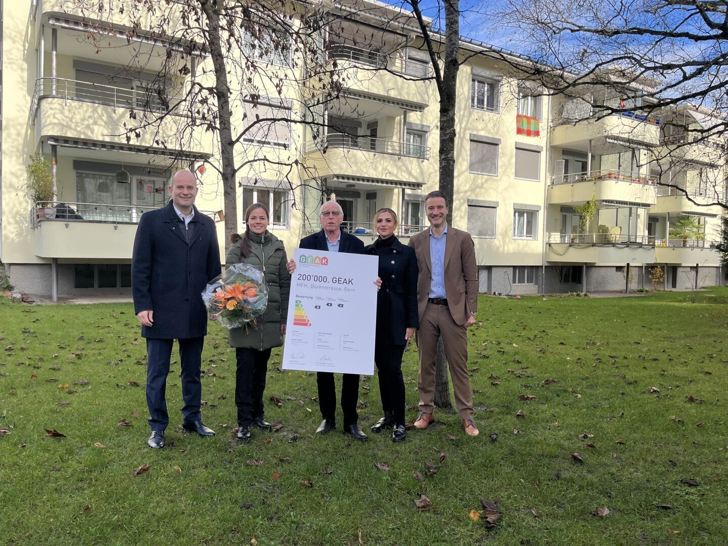 Fünf Personen stehen auf einer Wiese vor einem Wohnhaus. Drei von ihnen halten eine große Urkunde, eine Person einen Blumenstrauß in der Hand. Im Hintergrund sind Bäume ohne Blätter zu sehen.