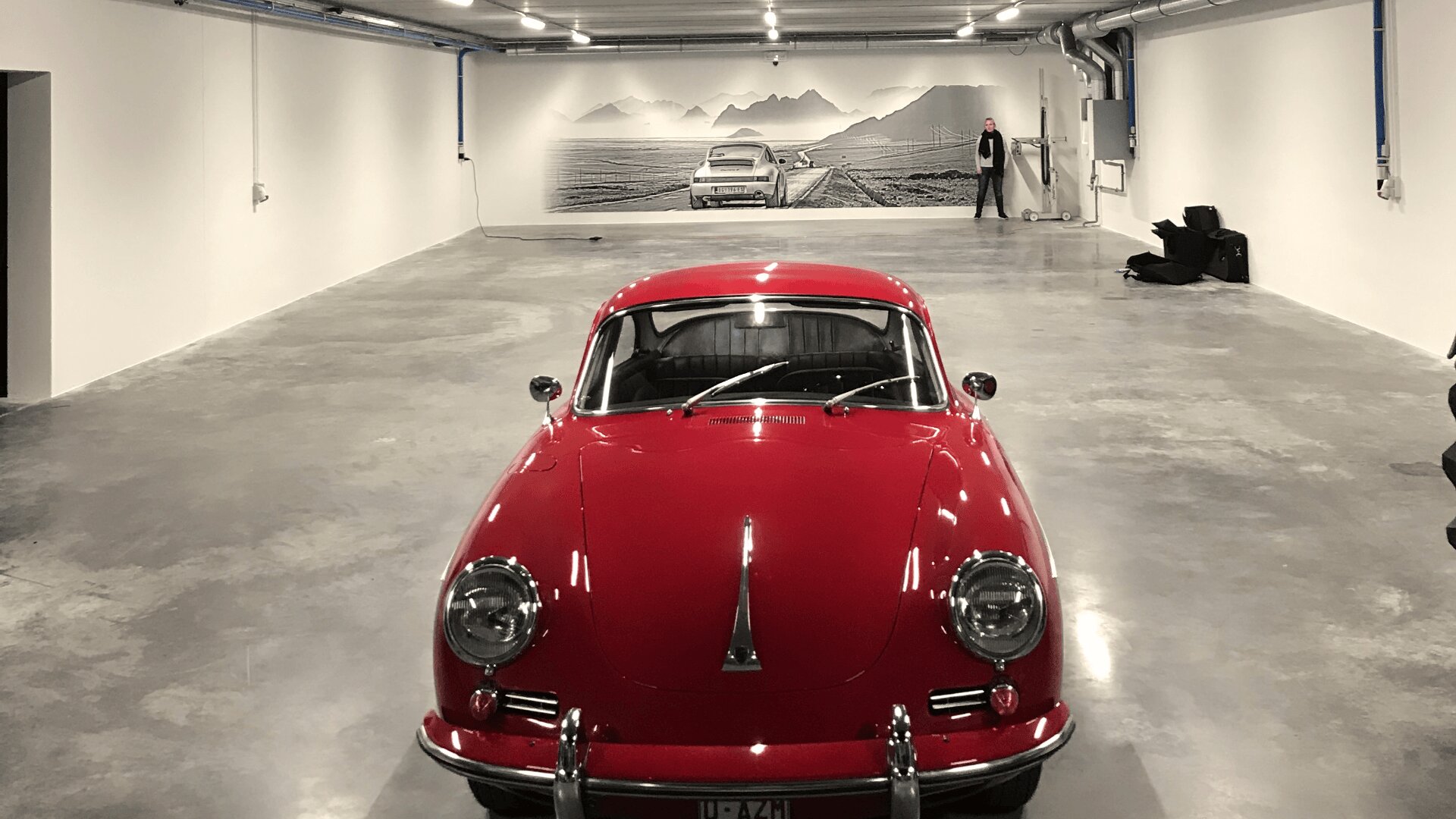 Une voiture de collection rouge et brillante est garée dans une grande pièce vide aux murs blancs et au sol en béton. À l'arrière-plan, une personne se tient près d'une peinture murale représentant une voiture de collection sur une route panoramique bordée de montagnes.