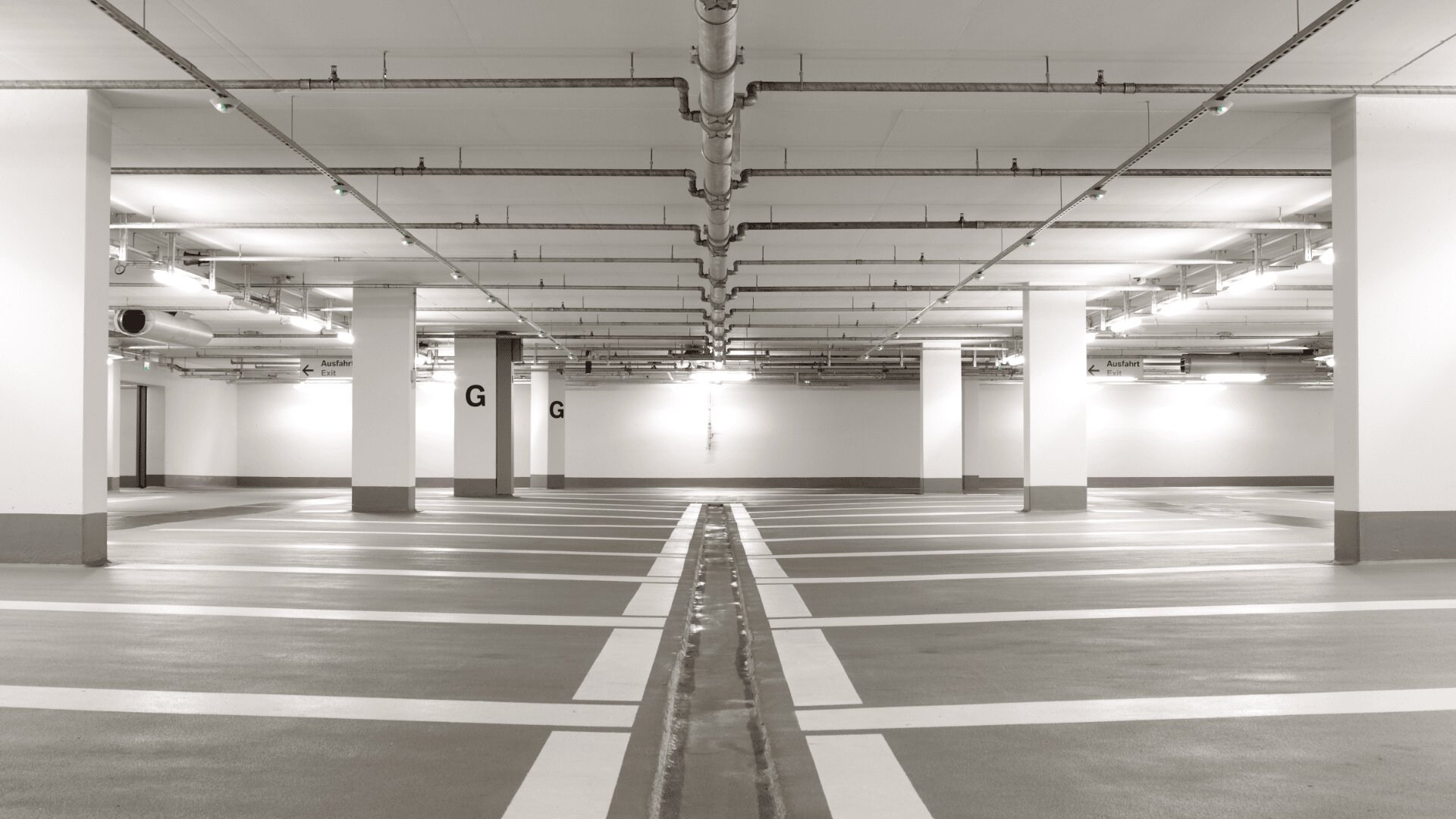 Leeres, gut beleuchtetes Parkhaus mit weißen Wänden, grauem Boden und weiß gestreiften Parkplätzen. Die Decke wird von mehreren weißen Säulen gestützt, und an der Decke darüber befinden sich grüne Anzeigeleuchten.